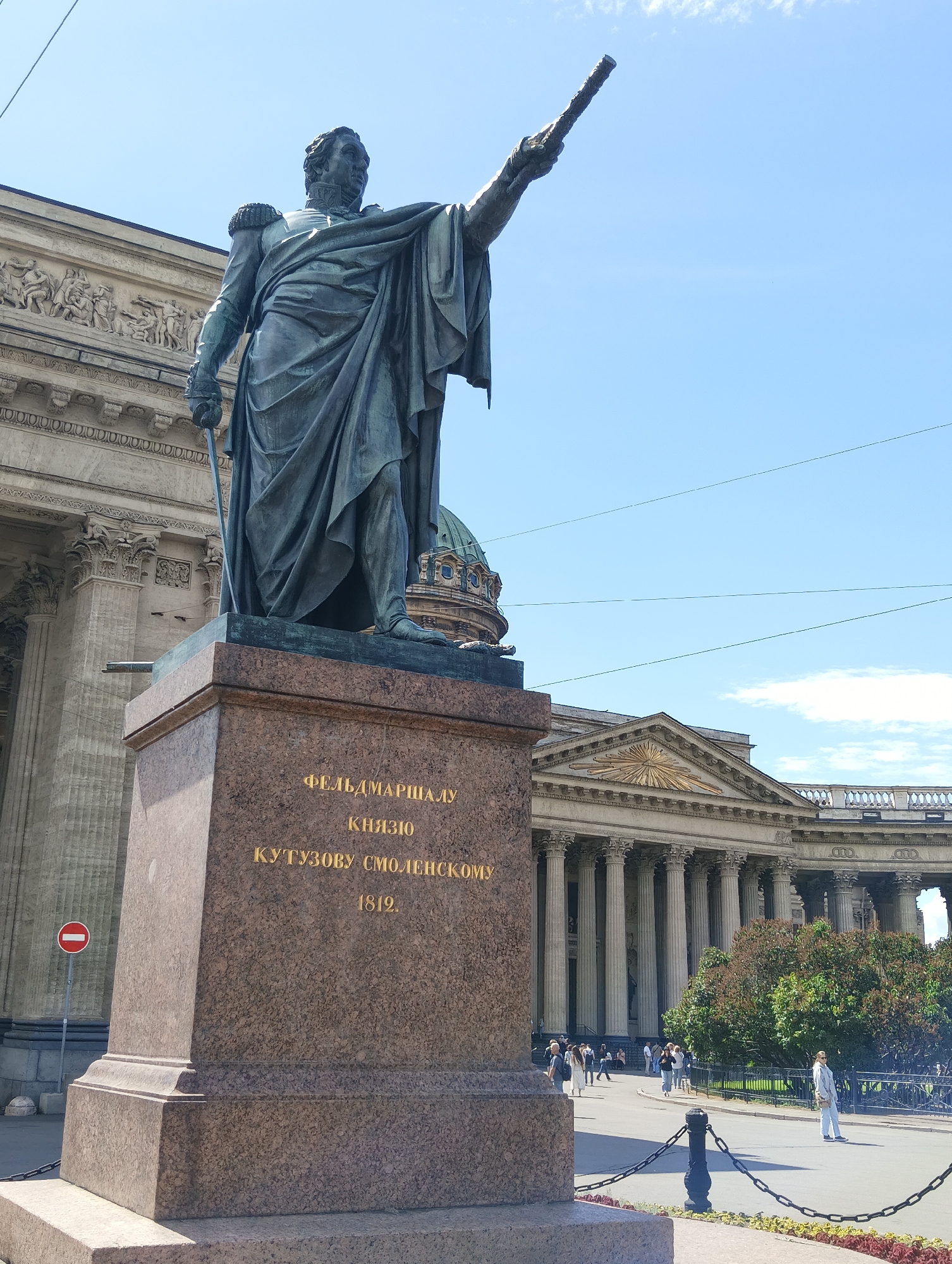 This media file features a bronze statue of a man in military attire, likely a general, with one arm extended upwards, holding a baton. The statue stands on a granite pedestal inscribed with text in Cyrillic. The text on the pedestal reads "ФЕЛЬДМАРШАЛУ КНЯЗЮ КУТУЗОВУ СМОЛЕНСКОМУ 1812", indicating it commemorates Field Marshal Prince Kutuzov of Smolensk and the year 1812.
The statue is positioned in front of a grand neoclassical building with a colonnade and a dome visible in the background. This architectural style, along with the inscription, suggests the location is Saint Petersburg, Russia, specifically in front of the Kazan Cathedral. The scene appears to be set on a clear, sunny day with a bright blue sky. A few wispy clouds are present.
Several people are visible in the background, strolling around the plaza and walking on the paved area in front of the building. They are dressed in casual summer clothing. A red and white circular "no entry" sign is placed to the left of the statue's pedestal. Black metal chains and posts delineate an area in the foreground, possibly a garden bed with flowers, though it is partially out of focus. The overall atmosphere is one of a public square on a pleasant day, with historical significance implied by the prominent statue and architecture.