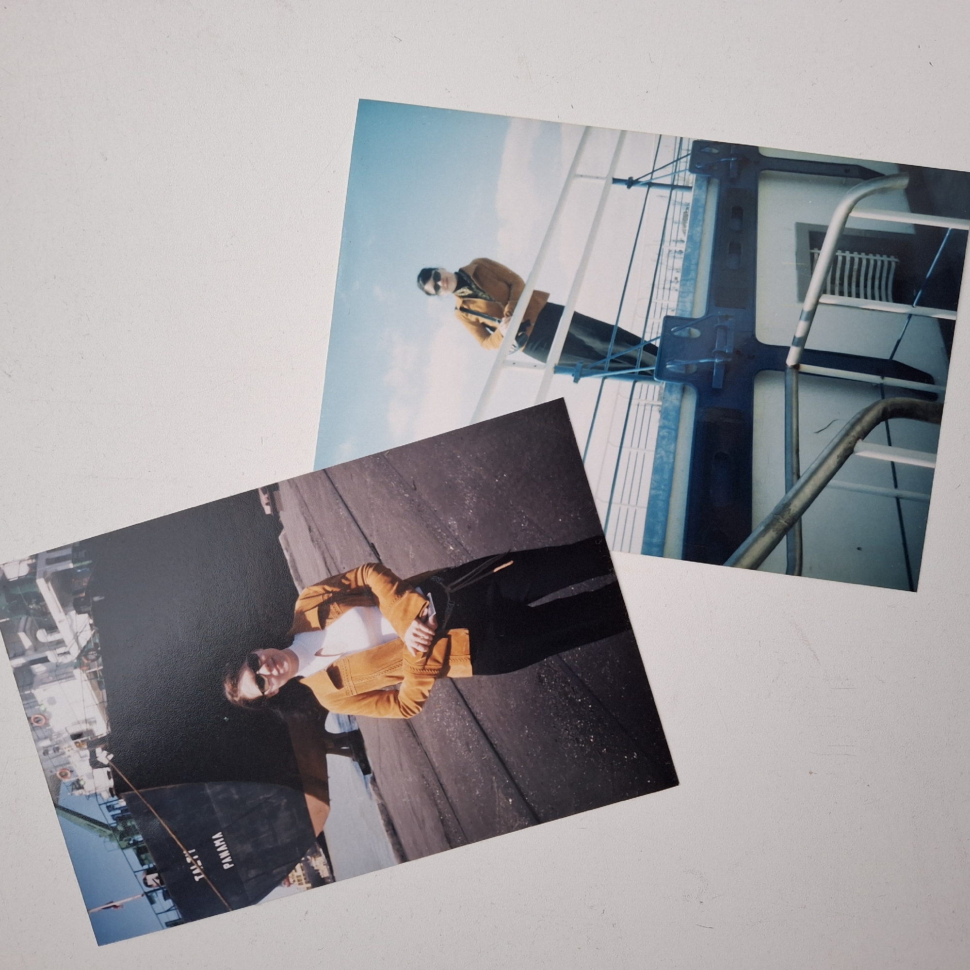 The image shows two photographs placed on a white surface. The photograph on the bottom left appears to depict a person standing on a pier beside a ship named "TALLI PANAMA". The person is wearing sunglasses, a white shirt, and a brown jacket with black pants. The photograph on the top right shows the same person wearing sunglasses and a similar outfit, leaning against a ship's railing with a blue sky in the background. The photographs seem to be taken on a ship or near the port, potentially in an unknown city. The environment includes a ship, a pier, and the sky.