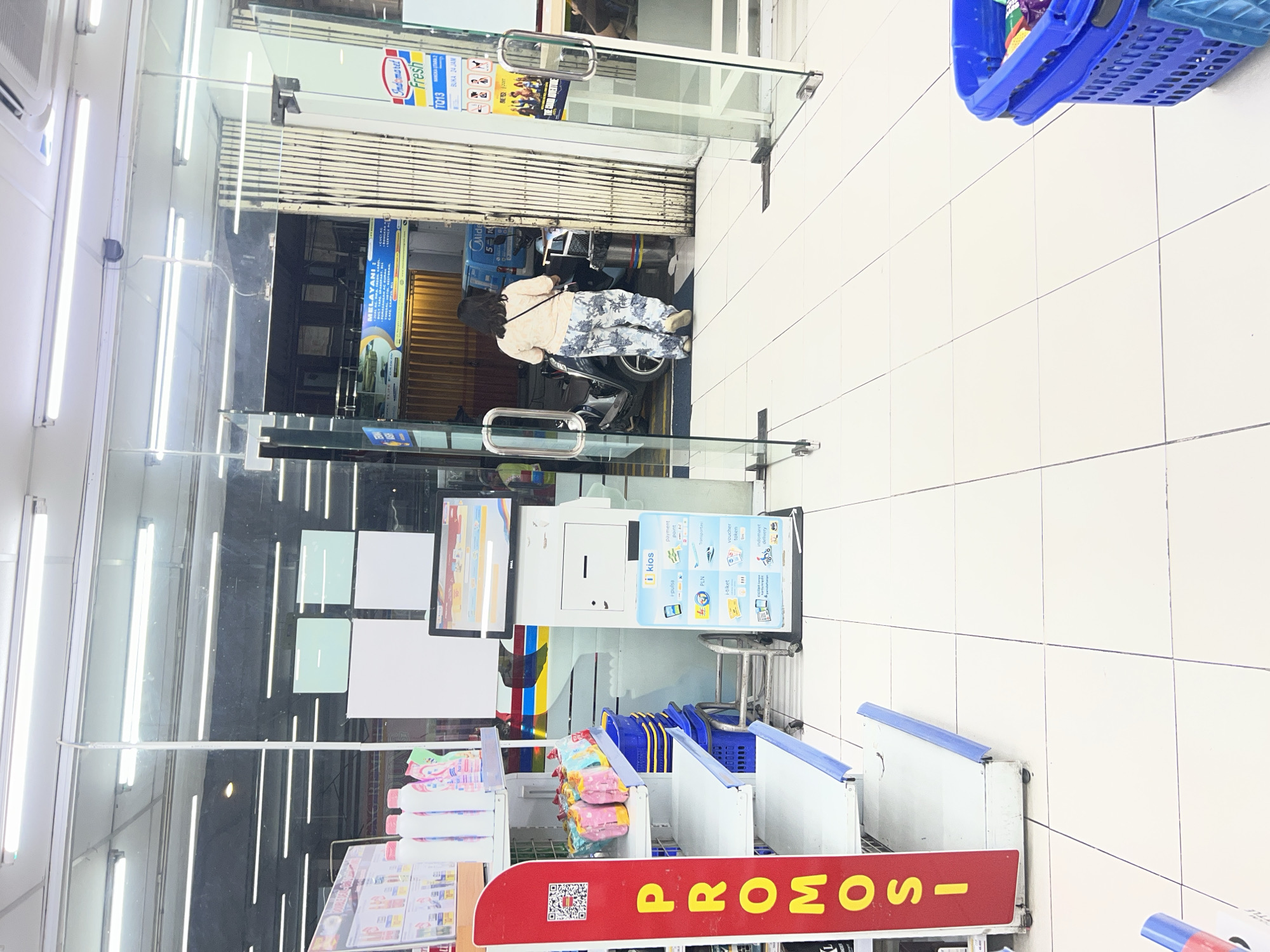 The image depicts the interior of an Indomaret convenience store in Denpasar, Indonesia, viewed from the entrance. The floor is tiled in a light beige color with dark grout lines. To the left, a red promotional stand with the word "PROMO" written vertically in yellow letters is visible, featuring a QR code. Behind the stand, shelves display several bottles of cleaning products, and lower shelves hold blue plastic baskets. In the center, a self-service payment kiosk, branded "i kios," is positioned near the glass doors. The kiosk has a screen displaying various services like i-pulsa, i-tiket, and PLN. A metal trolley with several blue baskets is also present.
Through the glass doors, a woman wearing a peach-colored top and patterned trousers walks away from the camera, heading towards a scooter parked in what appears to be a service or loading area. A large, collapsible metal gate is visible behind her. A sign for "Indomaret Fresh" is mounted on the right side of the gate, along with store information indicating it is open 24 hours. The store is illuminated by bright fluorescent lights on the ceiling. A blue shopping basket filled with items is partially visible in the lower right corner of the image.