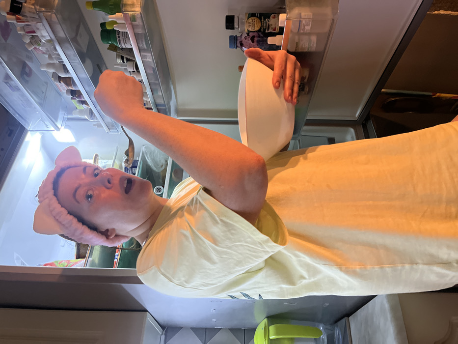 A woman wearing a pink headband with cat ears stands in front of an open refrigerator, holding a white bowl. Her facial expression indicates surprise, with wide eyes and an open mouth. The refrigerator's interior is lit, revealing shelves filled with various food items and condiments. The woman is wearing a yellow t-shirt. A green and yellow object sits on the counter below. The background shows a tiled wall.
