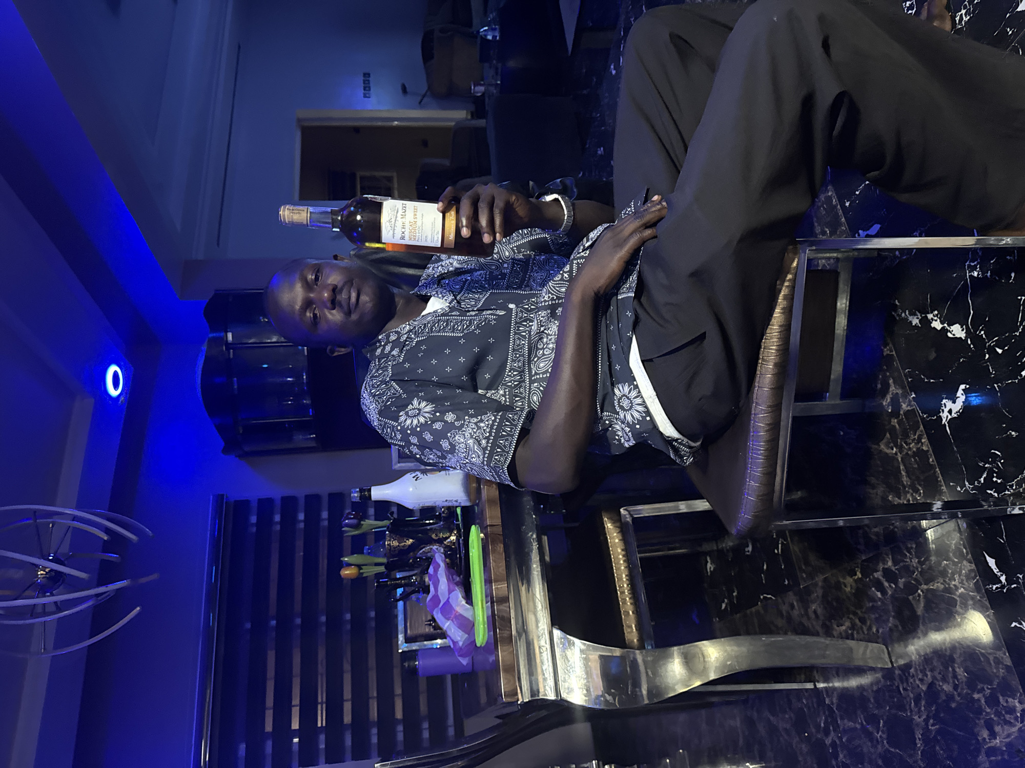 A man sits on a chair holding a bottle of "ROCHET MAZET Muscat de Beaumes de Venise Vin Doux Naturel" wine. The man is wearing dark trousers and a patterned shirt. The chair has a metallic frame and a leather seat. A bar-like structure is behind the man, adorned with bottles and decorations. The floor is a dark, marbled pattern. A blue light fixture is seen on the left side of the image. The location appears to be a well-lit, possibly residential space in the city of Unknown, Unknown.