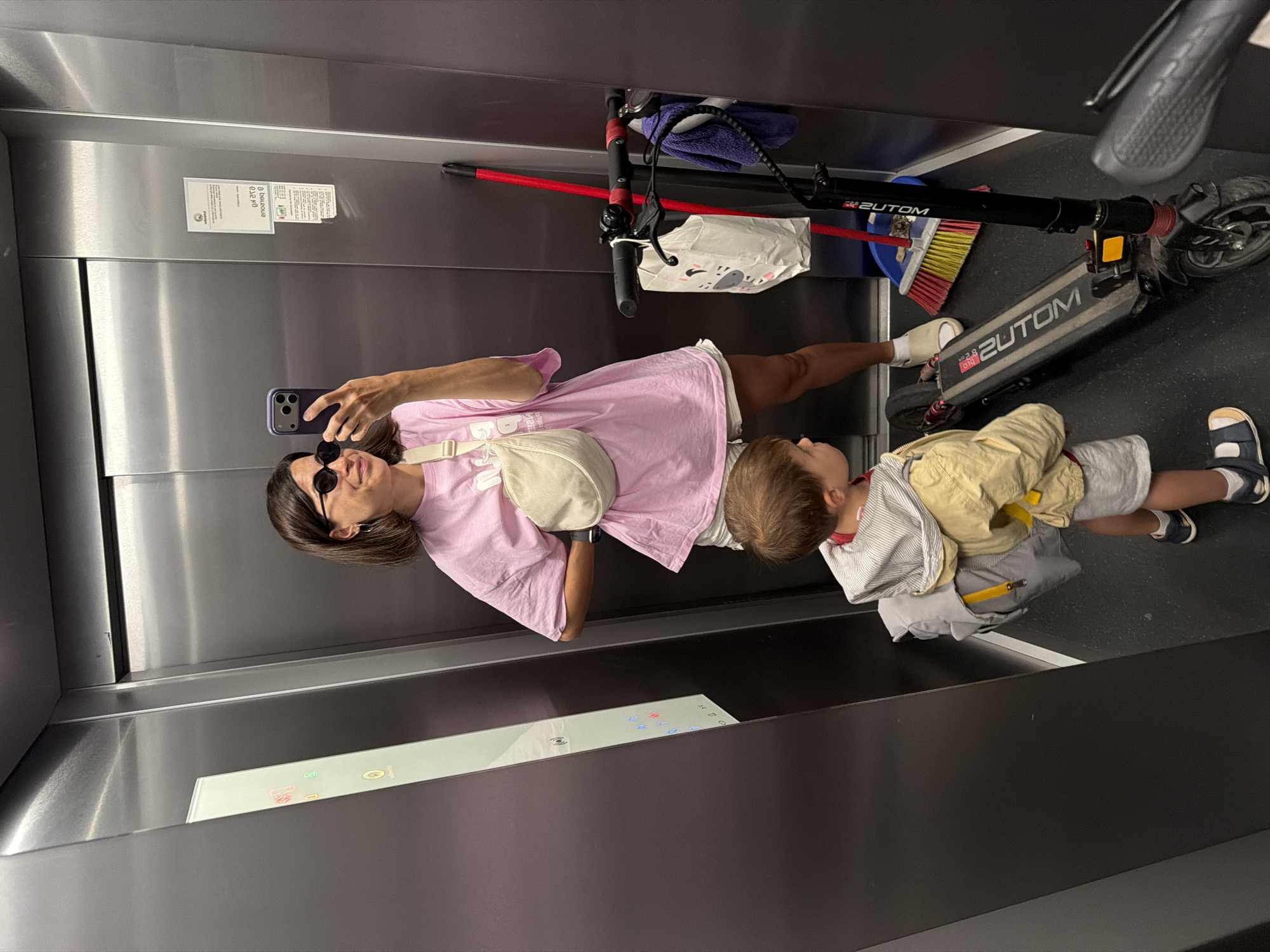 The photo was taken indoors, presumably in an elevator. It features a woman and a young child alongside a black scooter. The woman, wearing sunglasses and a light pink shirt, is holding a smartphone and appears to be taking a selfie. The child, wearing a yellow jacket and gray shorts, stands near the woman. The black scooter, labeled "2UTOM," is also present in the frame, along with a red broomstick. The elevator has stainless steel walls and a panel of buttons. Based on the presence of the 2UTOM scooter and other factors, this image was likely taken in Budva, Montenegro.
