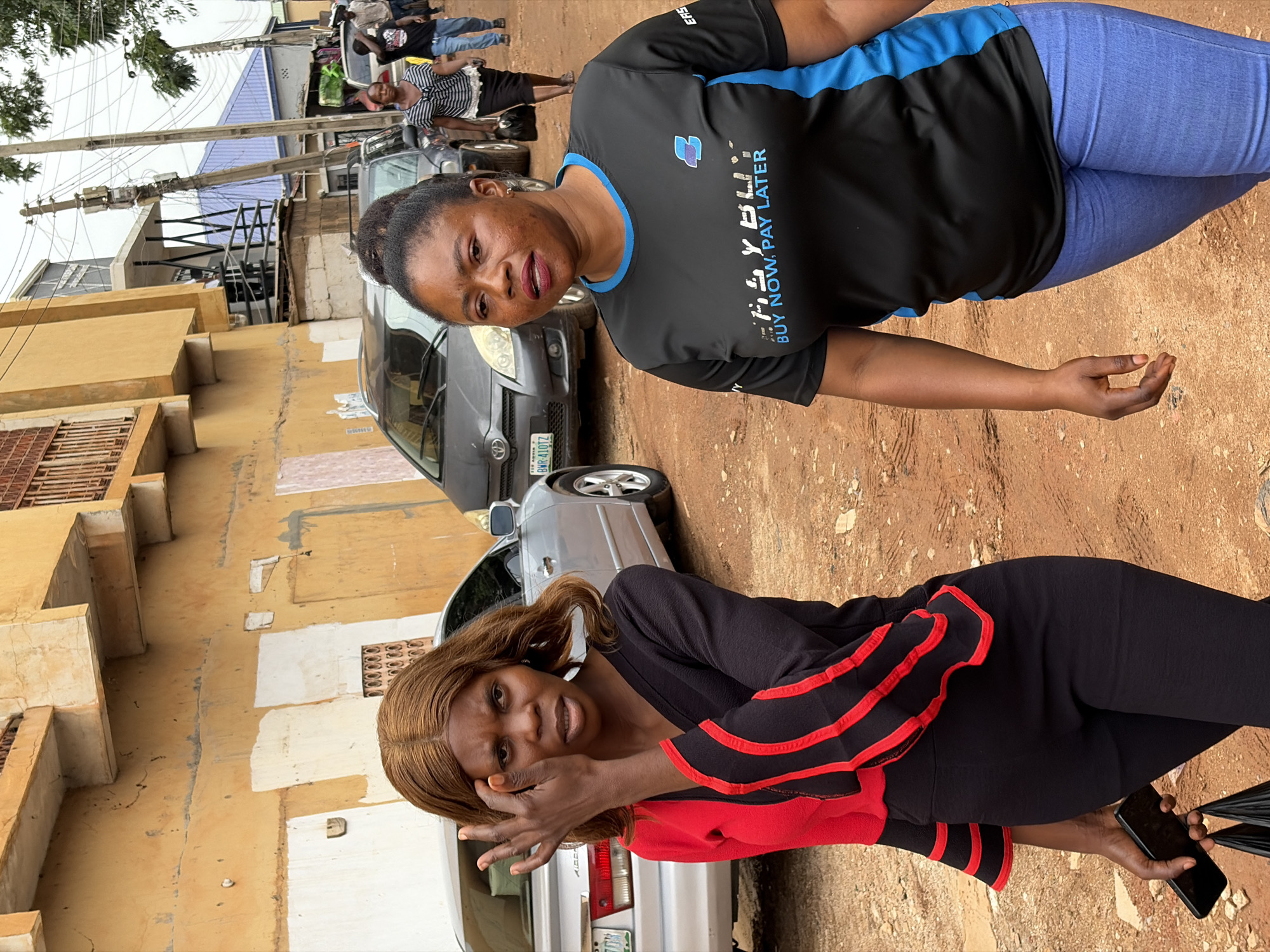 The image shows two women standing outdoors in Jos, Nigeria. One woman wears a black shirt with blue trim and blue pants; the shirt has text "Fly Buy" printed on it. The other woman wears a black and red top and black pants, holding a phone. They are positioned on a dirt area near a street with parked cars and pedestrian traffic in the background. A yellow building with window grilles is visible to the left. Utility poles and power lines are present.