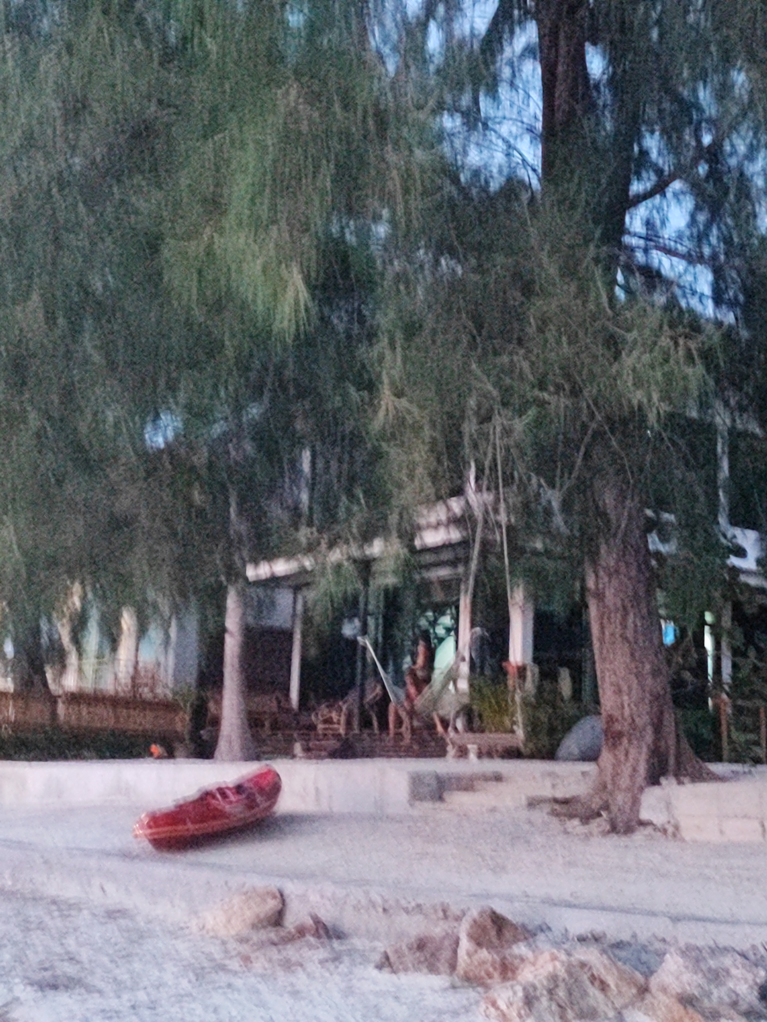 The image captures a scene on a beach in Ko Pha-ngan District, Thailand. In the foreground, a red kayak rests on the sandy shore, angled slightly to the right. Beyond the kayak, a pathway or concrete structure runs horizontally, bordered by sand. Further back, lush green trees with dense foliage dominate the middle ground, with a large tree trunk visible on the right side. Behind the trees, a building with a white roof and visible pillars suggests a structure like a cafe or resort. A person can be seen seated on a swing or hammock beneath the trees. The overall environment suggests a tropical beach setting, likely in the late afternoon or early evening due to the soft lighting. The image is slightly blurred, giving it a somewhat abstract quality.
