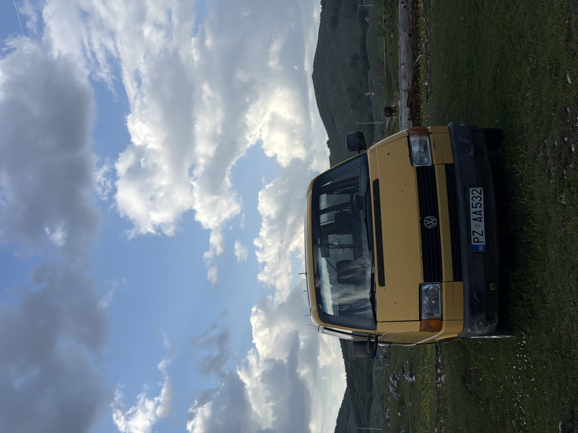 The image depicts a yellow Volkswagen van parked on a grassy field in Trsa, Montenegro. The vehicle's license plate reads "PZ AA532." The background features a mountainous landscape with a cloudy sky. A fence or wooden barrier can be seen in the distance. A dark-colored animal, possibly a horse or cow, is visible behind the van near the fence. The van is angled slightly towards the viewer. The grass appears to be dotted with small white flowers.