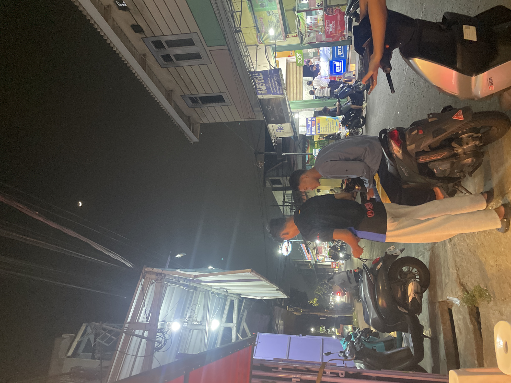 The image depicts a street scene at night, likely in North Jakarta, Indonesia. Two young men are prominently featured in the foreground. The man on the left is wearing a black t-shirt with blue and white accents and light-colored pants. He appears to be standing and looking towards the right. The man on the right is wearing a light gray long-sleeved shirt and dark pants, seated on a dark-colored scooter. He is looking forward, with his right hand resting on the handlebars. Another scooter, light blue, is parked to the left of the dark scooter.
The background reveals a street lined with shops and businesses. Signs with Indonesian text are visible, including one for "MARS CELL" and another mentioning "JUICE" and "MARTABAK". Several other scooters and a few people are present on the street, some riding and others standing. The buildings are of a modest size, with overhead wires and lighting contributing to the urban nighttime atmosphere. A sliver of a crescent moon is visible in the dark night sky. The overall impression is of a busy, casual street environment at night.