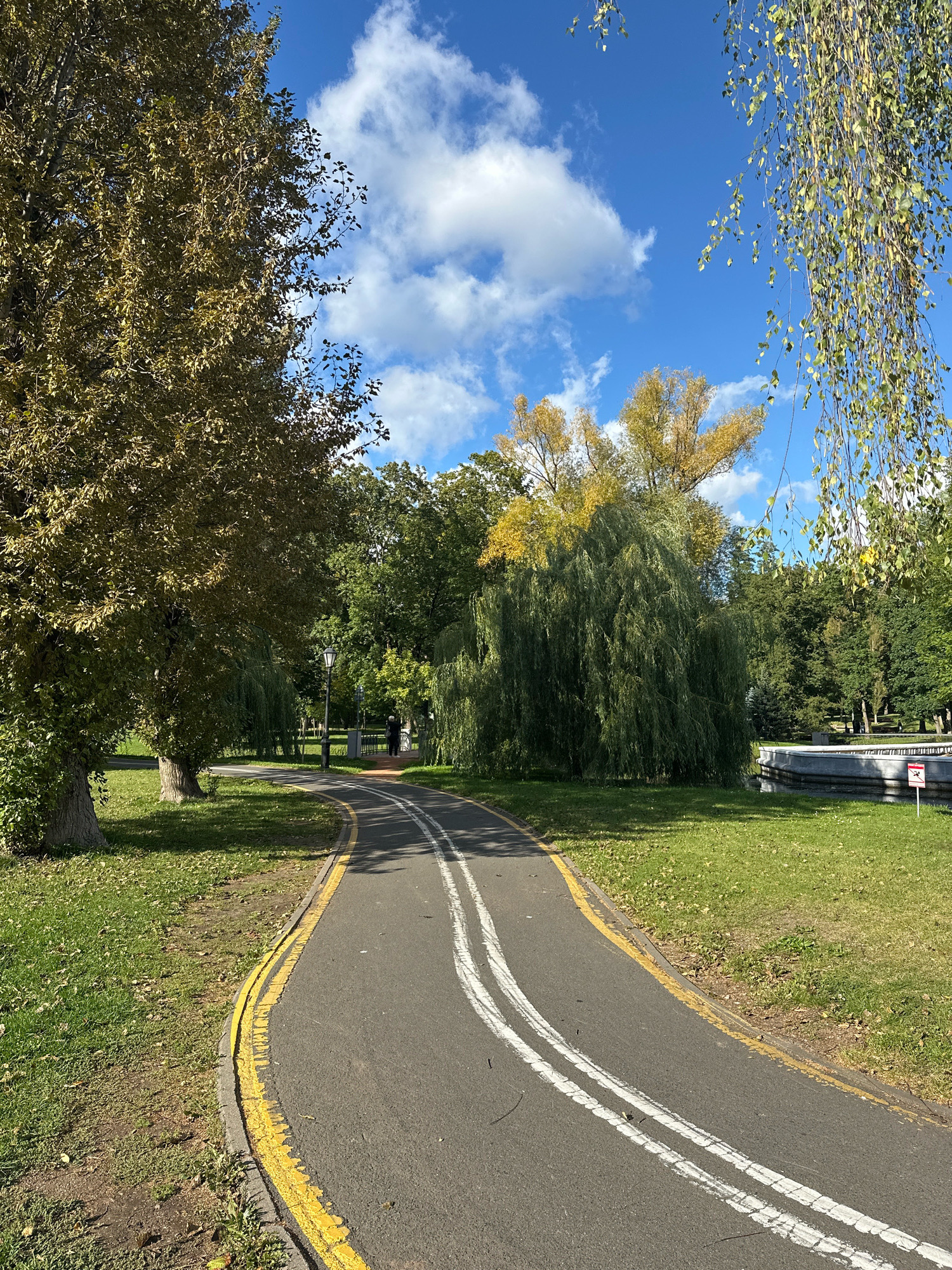 The image depicts a paved pathway with yellow and white markings in a park setting under a bright blue sky with scattered clouds. The pathway curves to the right, bordered by green grass and trees. On the left, a large tree with brown and green foliage stands prominently. Further down the path, a lamppost is visible, and a person dressed in dark clothing is walking away from the camera. In the background, there are more trees, including a weeping willow with green foliage. To the right, there is a body of water with a light-colored edge and a sign with red markings. The overall scene suggests an autumn day in a park. This image was taken in Minsk, Belarus.