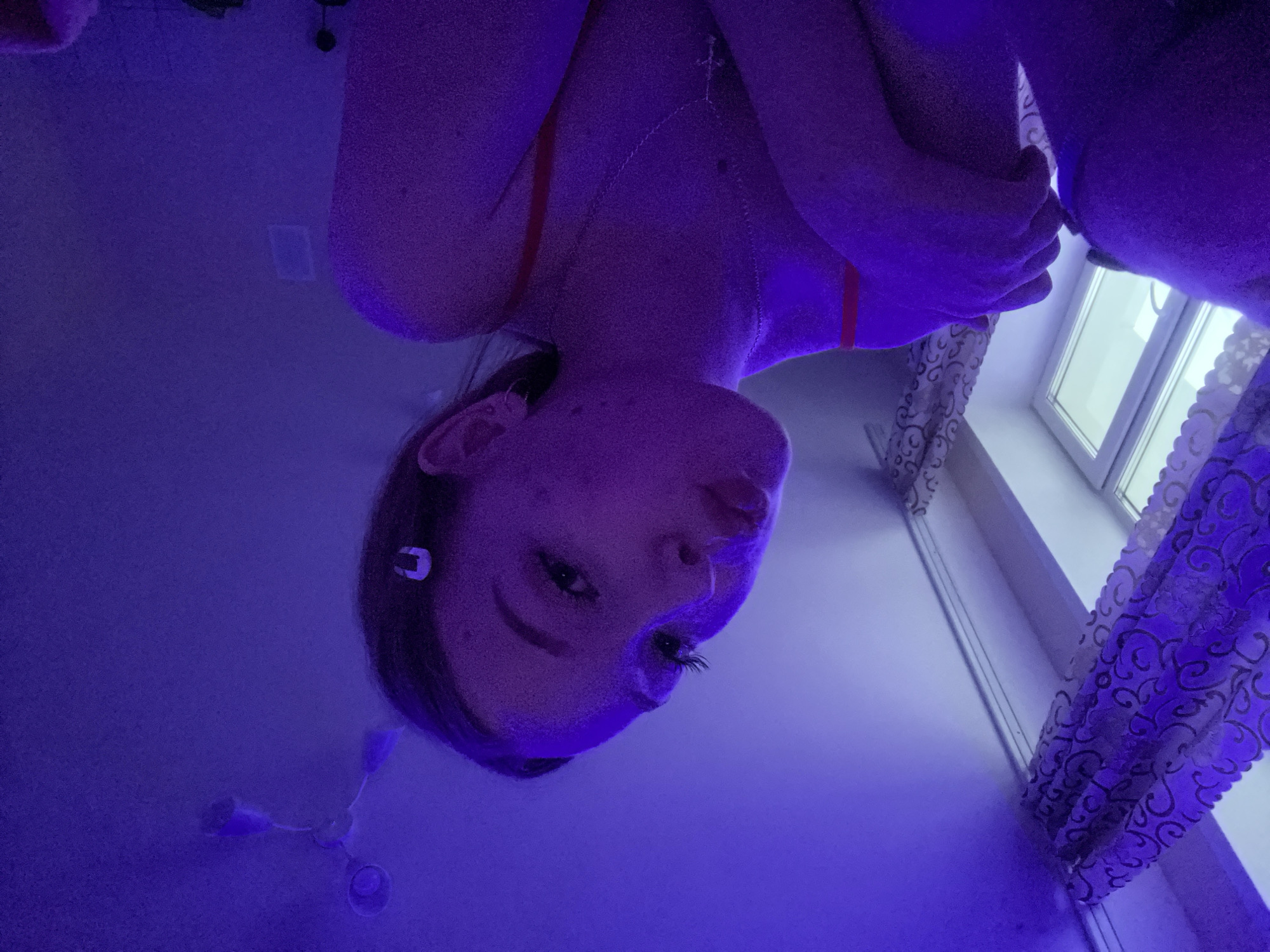 A selfie of a woman taken from a low angle in a room with purple lighting. The woman has fair skin, dark hair, and wears a small earring. She also wears a silver necklace with a cross. Her arms are crossed. In the background, there's a window with patterned curtains. A ceiling fan and a light switch are also visible on the wall. The location is possibly in a residential setting. The city of Chaikovsky, Russia, cannot be definitively confirmed from the visual information provided.