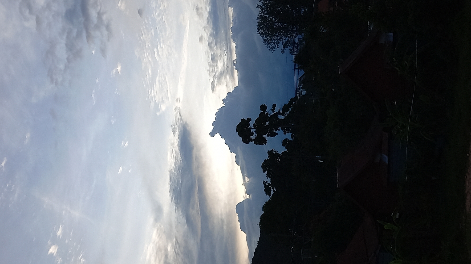 The image depicts a silhouette of a tropical landscape at dusk or dawn, likely in the Ko Pha-ngan District of Thailand. The foreground is dominated by the dark outlines of several houses with reddish-brown, sloping roofs, partially obscured by dense foliage. Beyond the houses, a lush, dark green tree line rises, transitioning into hills on the left side of the frame. A solitary, tall tree with a distinctive silhouette stands prominently in the mid-ground, its branches reaching towards the sky. The sky itself is a dramatic canvas of clouds, with a bright, hazy light source peeking through a gap in the heavy, dark clouds in the upper center. These clouds are sculpted into dramatic shapes, suggesting either an approaching storm or the aftermath of one. The overall mood is serene yet dramatic, characteristic of twilight in a tropical region. There are no visible people or animals.