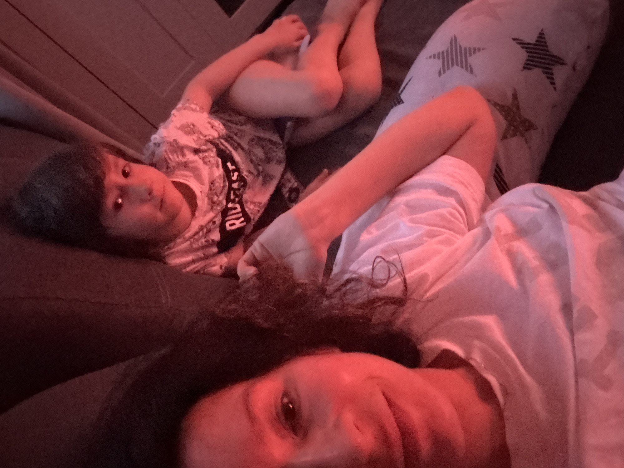 A selfie-style photograph shows a woman and a child lying on a sofa in what appears to be an indoor setting in Chaikovsky, Russia. The woman, positioned at the bottom of the frame, looks at the camera with a partial smile. Her hair is dark and curly. A white t-shirt covers part of her body. The child, lying behind and to the left of the woman, is wearing a patterned t-shirt with the word "RIDE" visible. The child is also looking at the camera. Their legs are visible near the top of the frame, positioned to the left. A pillow with star patterns is to the right of the child. The lighting in the room casts a reddish-pink hue on everything. The sofa is dark. A portion of a wall is visible in the upper left corner.