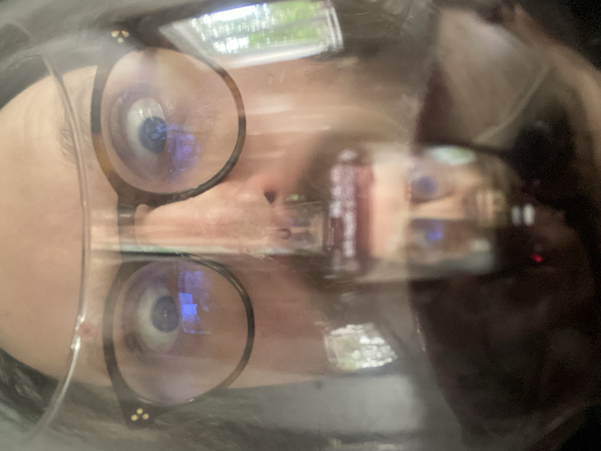 The image shows a close-up of a face with glasses, likely taken from a distorted perspective. The reflection in the glasses suggests an outdoor environment with greenery and structures, possibly in Rusanovo, Russia. The details are obscured by the reflection and distortion. The person is wearing eyeglasses and the image is of their face, partially distorted due to reflections.