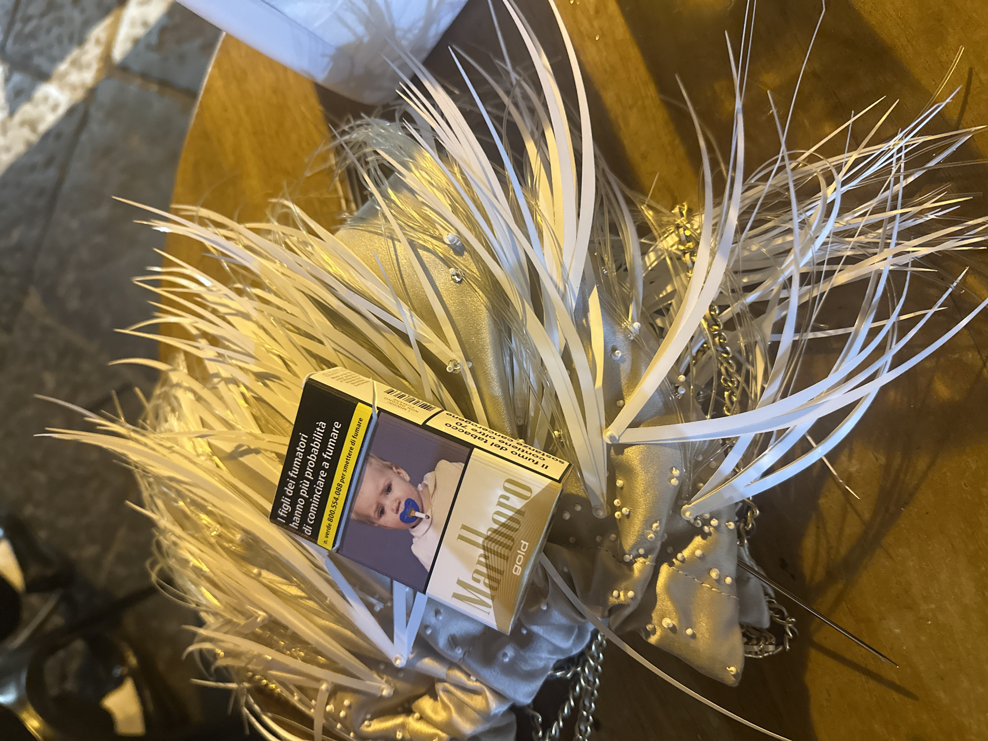 Here is a description of the image:
The image presents a close-up view of a pack of Marlboro Gold cigarettes placed on top of a decorative handbag or clutch. The pack of cigarettes features a health warning in Italian, including an image of a baby with a pacifier. The text indicates the children of smokers are more likely to start smoking, including a number to help quit smoking. The handbag is light in color and adorned with numerous white plastic strips or feathers, giving it a feathered or textured appearance. There are also small, shiny embellishments scattered across the bag, possibly beads or rhinestones. The overall setting appears to be indoors, with a dark background or surface below. The lighting is dim, casting soft shadows. It's possible the pack of cigarettes and handbag are being presented as a still life or casually placed together.