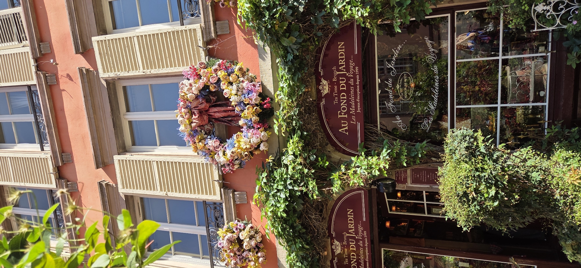 The image depicts a charming facade of a shop, likely in Strasbourg, France. The building features a salmon-pink exterior with multiple windows framed by beige wooden shutters. Large, circular floral wreaths, one prominently displayed, adorn the wall, adding a festive and welcoming touch. Lush green ivy cascades down the building, framing the entrance and windows.
A prominent sign with a burgundy background and gold lettering reads "AU FOND DU JARDIN" and "Les Madeleines du Voyage... Joyaux d'exception depuis 1999". The reflection in the shop window also shows the name "Au Fond du Jardin" and "MAISON DE GOÛT", suggesting it is a specialty food shop or tearoom. The presence of the floral wreaths and the shop's name evoke a sense of tradition and perhaps a celebration. The lighting suggests it is daytime, with the sun casting shadows, indicating fair weather. The overall scene conveys a quaint and inviting atmosphere, characteristic of a European street.