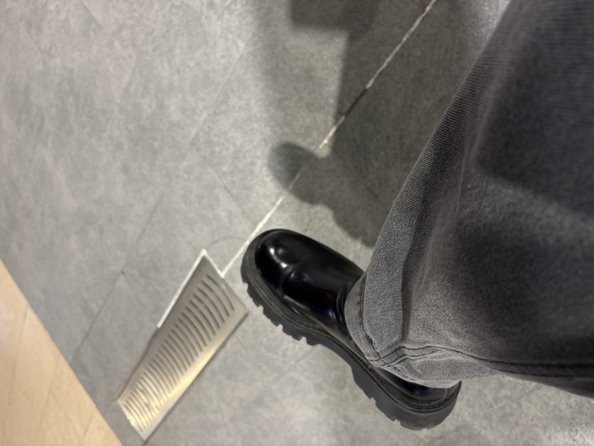 A single, black, shiny, leather boot with a thick, lugged sole is positioned on a gray tiled floor next to a narrow, rectangular, metal ventilation grate. The boot is mostly covered by a pair of gray denim jeans. The location is inferred to be in the city of Puebla, Mexico.