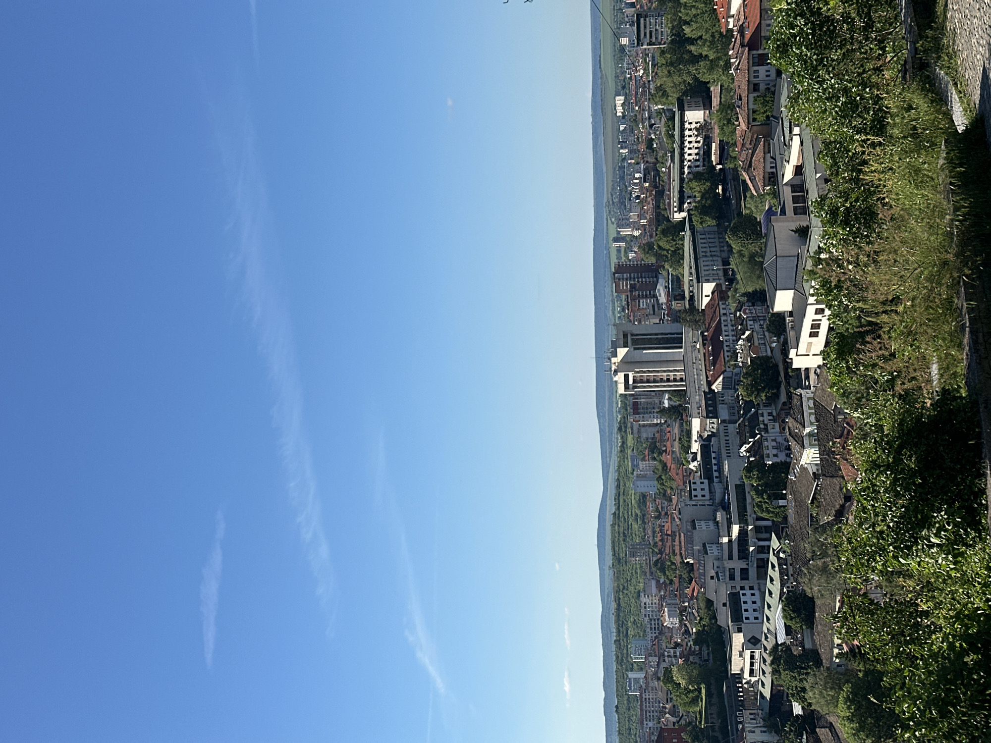 The image shows a cityscape under a blue sky with scattered wispy clouds. The buildings are densely packed, and there is lush green vegetation along the edge of the frame. The scene could be Lovech, Bulgaria, a city known for its elevated views and architecture.