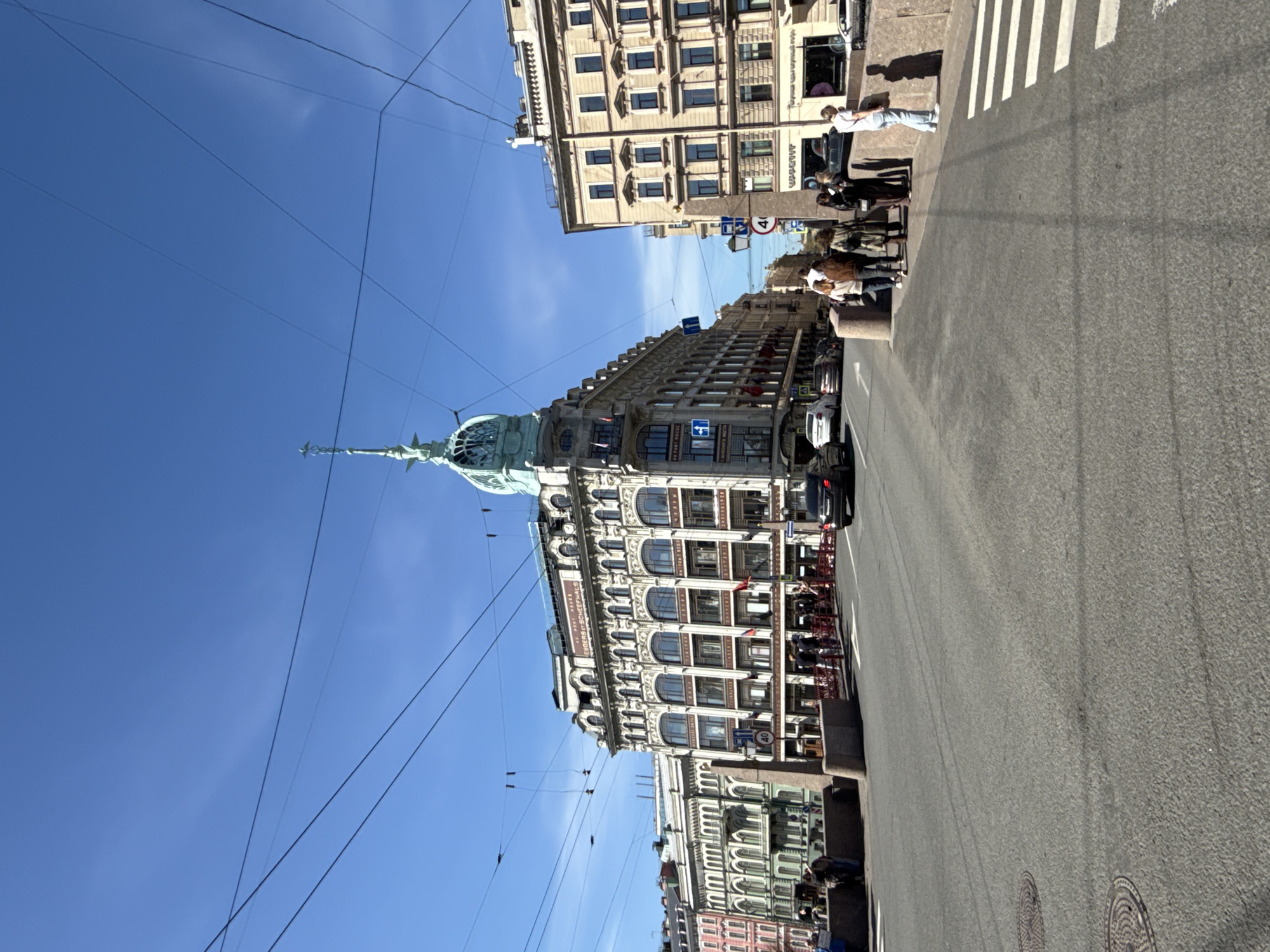 The image captures a street scene in Saint Petersburg, Russia, under a clear blue sky intersected by numerous overhead power lines. The focal point is a tall, ornate building with a distinctive turret and spire, indicative of historic architecture. Adjacent to it, another multi-story building with classical window designs is visible. Pedestrians are seen walking along the sidewalk and crosswalk, and a few cars are present on the asphalt road. Restaurant seating is visible.