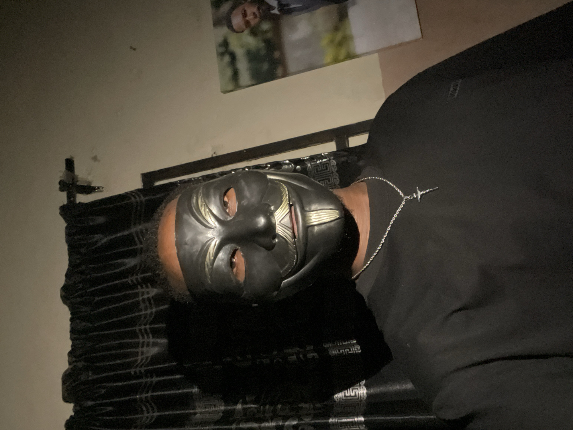 The image shows a person lying down, possibly on a bed, wearing a black mask. The person is also wearing a black top and a silver necklace with a cross pendant. A framed picture hangs on the wall above the head of the bed. The location is unknown, and no indication of Lokoja, Nigeria is present.