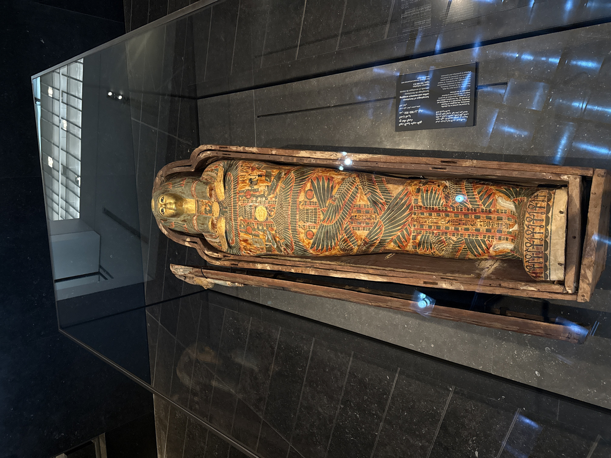 This image shows an ancient Egyptian sarcophagus, likely on display in a museum. The sarcophagus is elaborately decorated with colorful hieroglyphs and depictions of deities and mythological scenes. The golden mask of the mummy is visible at the top. The sarcophagus is housed in a wooden outer coffin, which is partially open. The display is illuminated by artificial lighting, creating reflections on the glass casing and surrounding surfaces. A descriptive plaque is visible to the right of the sarcophagus. The background appears to be a modern architectural space with dark stone flooring and reflective walls or windows, suggesting an indoor exhibition setting. The scene is forensic and detailed, capturing the artifact and its presentation within a controlled environment. The location of this artifact in Abu Dhabi is the Louvre Abu Dhabi.