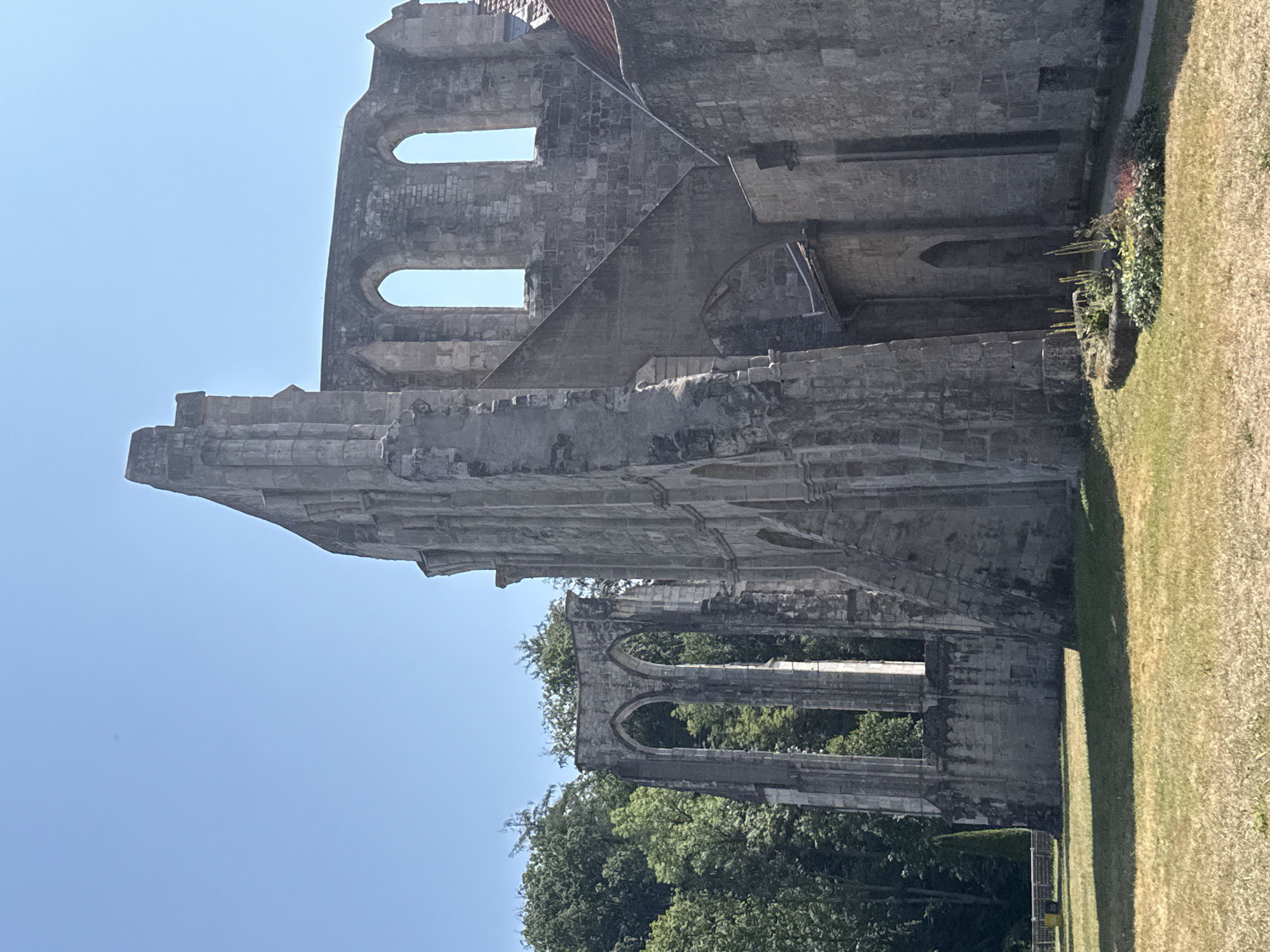In Walkenried, Germany, the image captures the ruins of a former Cistercian abbey, specifically the Gothic nave, under a clear blue sky. The stone structure, characterized by its weathered facade and arched openings, stands against a backdrop of lush green trees. The foreground features a sun-drenched grassy area with a small, manicured flowerbed. The scene is devoid of any human presence or activity.