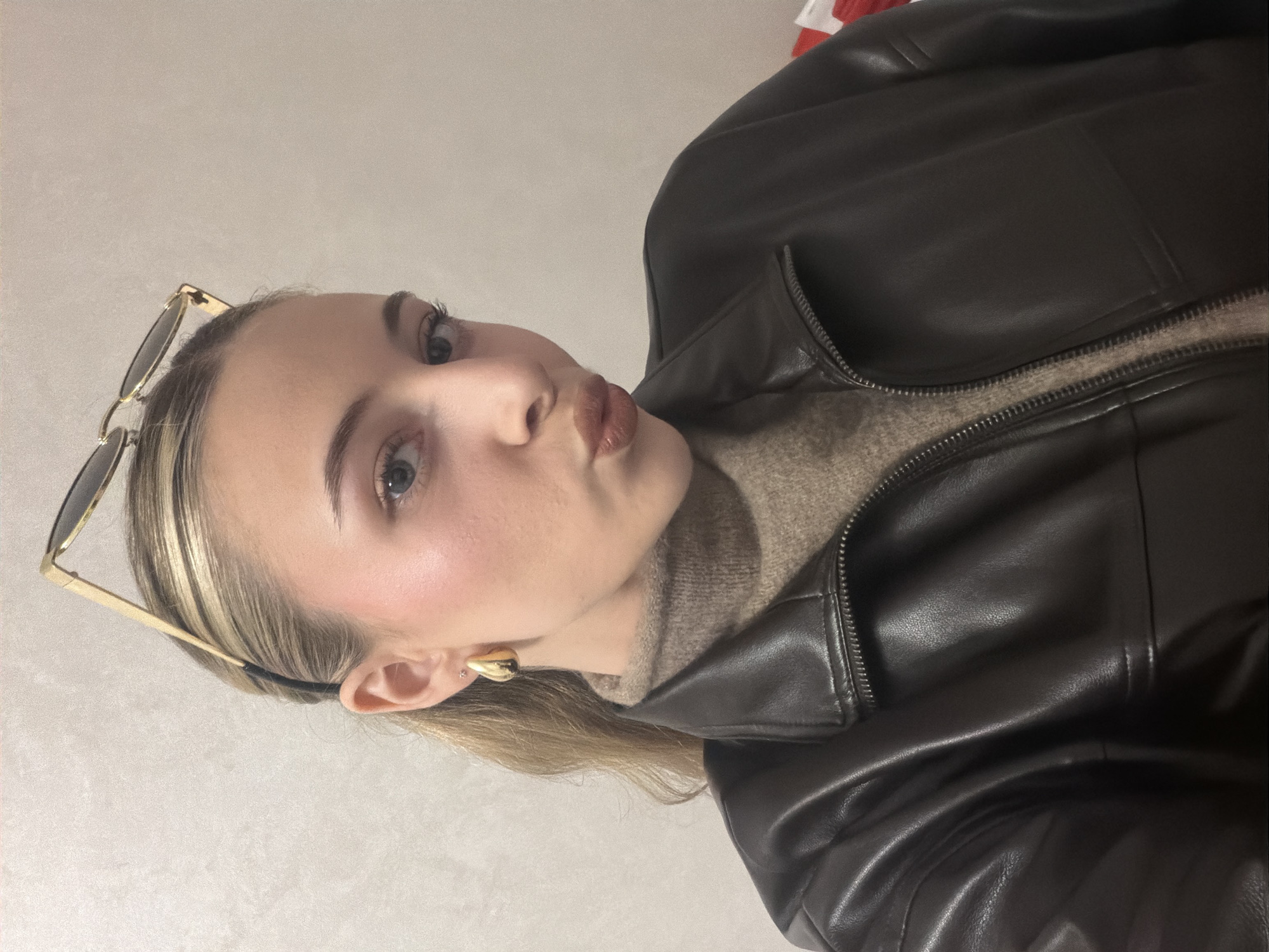 A close-up indoor photograph depicts a young female subject from the chest up, looking directly at the camera with puckered lips. She has light skin, blue eyes, and light-colored hair pulled back from her face, with gold-framed sunglasses resting on top of her head. She wears gold-toned teardrop-shaped earrings and a smaller stud earring in her earlobes. Her attire consists of a light brown or taupe knitted turtleneck sweater layered under a dark brown, zippered faux leather jacket, which is partially unzipped. The background is a plain, light-colored wall with a subtle textured finish. No specific visual information in the image indicates the location in Plovdiv, Bulgaria.