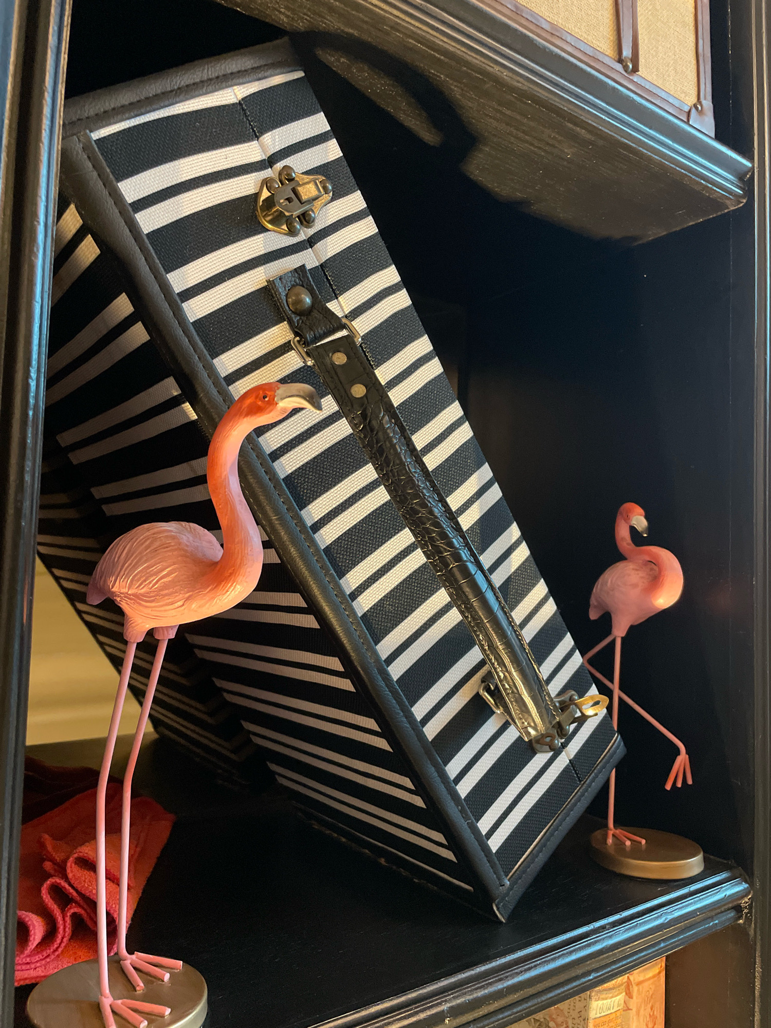 This is a close-up, eye-level, indoor photograph taken at a slight upward angle. The scene appears to be a shelf within a dark wooden cabinet or bookcase. The primary subjects are two pink flamingo figurines and a black and white striped suitcase.
The flamingo on the left is larger and positioned in the foreground. It is standing on thin pink legs attached to a circular, metallic base. Its neck is curved upwards and to the right, with its head tilted slightly. The flamingo on the right is smaller and further back, also standing on thin pink legs attached to a similar base. Its neck is curved upwards and to the left, with its head tilted down.
The suitcase is leaning against the back of the shelf, tilted towards the viewer. It has a black and white striped pattern and a black leather handle with a textured finish. A brass-colored latch is visible on the front of the suitcase, and a black strap with rivets is attached to the side.
To the left of the flamingo in the foreground, a folded piece of red fabric, possibly a cloth or towel, is visible on the shelf.
The background is dark and out of focus, suggesting the interior of the cabinet. The shelf itself is dark wood with a subtle sheen, and its edge has a decorative molding. The overall impression is a whimsical and decorative arrangement.
There is no information in the image that would suggest the location is Pattaya City, Thailand.