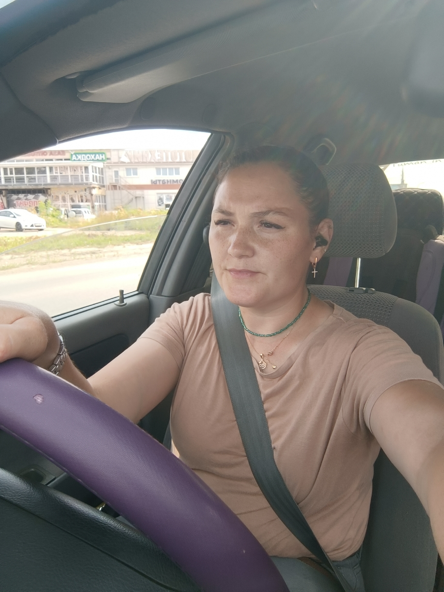 A woman is driving a car, holding the purple steering wheel with her left hand. She is wearing a tan t-shirt, a silver watch, and two necklaces. She has on a seatbelt that crosses her chest. In the background, there are buildings with signs that read "АЖДОХАН" and "ПТБИММО". The car is on a road with other cars visible. The environment appears to be a city street in Chaikovsky, Russia.
