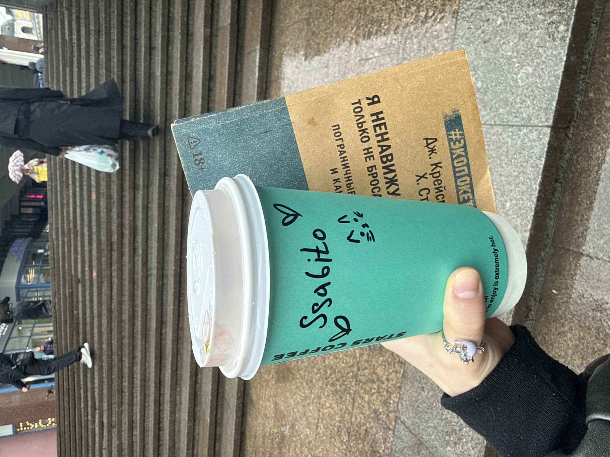 Here is a detailed description of the image:
**Overall Scene:** The image appears to be taken outdoors, likely in a city, during the day. The focus is on a hand holding a "Stars Coffee" cup with the name "Ssabito" written on it. There's also a book visible, with text in Russian on it. In the background, there are stairs and people walking.
**People:**
* A hand is holding the coffee cup. It has a ring on one of the fingers. The wrist is covered by a black sleeve, presumably of a jacket or sweater.
* In the background, people are walking on the stairs. One is wearing a black coat.
**Objects:**
* Coffee Cup: It's a teal-colored "Stars Coffee" cup with a white lid. The name "Ssabito" is written on it in black marker.
* Book: A book with Russian text is visible. The text includes the phrase "Я НЕНАВИЖУ" ("I hate") and a hashtag "#ЗКОПОКЕТ" (likely a shortened word related to ecology/environment) can also be seen.
* Stairs: Gray, concrete stairs are in the background.
* Clothing: A black coat, other clothing on background people.
* Ring: Visible on the hand holding the cup.
**Environment:**
* The setting seems to be an urban environment, possibly near a building with stairs leading up or down.
* It seems to be outdoors, with natural lighting.
**Possible Location in Moscow, Russia:** Given the brand of the coffee cup ("Stars Coffee", which replaced Starbucks in Russia), the Russian text on the book, and the style of architecture/environment in the background, it's reasonable to speculate that the photo was taken in Moscow, Russia.