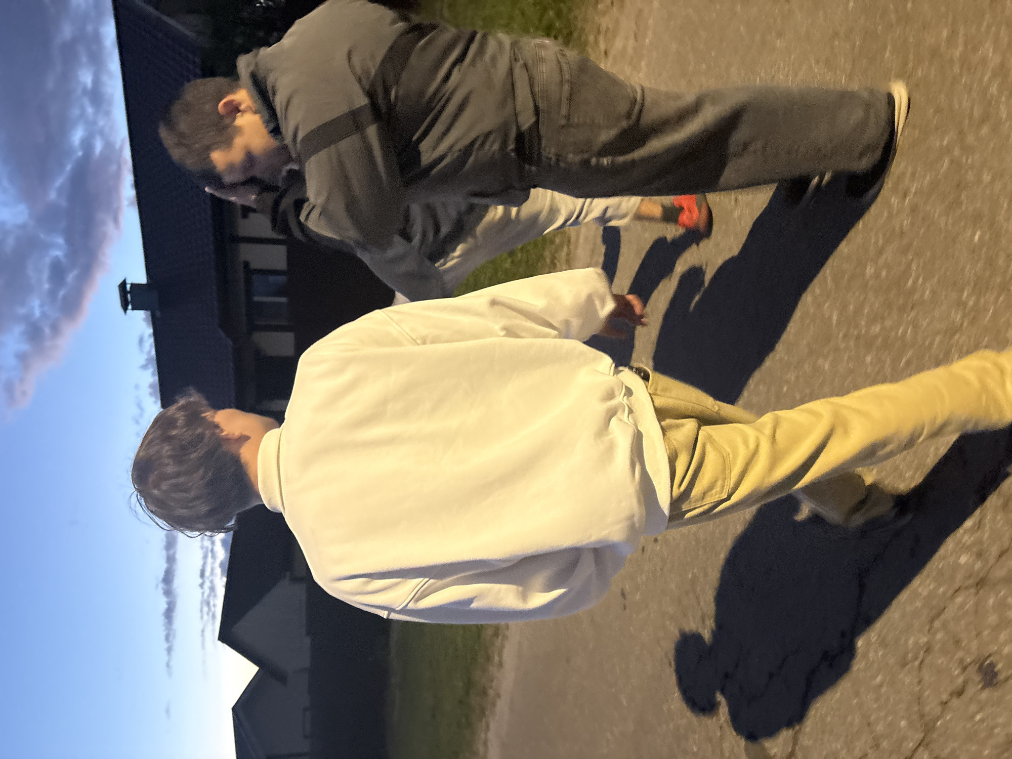 The image depicts three individuals outdoors, possibly walking or standing on a paved surface like a road or path. The scene is illuminated by either early morning or late evening light, indicated by the deep blue sky with scattered clouds and long, prominent shadows cast on the ground.
In the foreground, with their back to the camera, is a person wearing a light-colored (possibly white or off-white) jacket and light yellow or khaki pants. This individual has short, dark hair. Their shadow stretches out long in front of them, suggesting a low light source, likely the sun during twilight hours.
Behind and slightly to the left of the first person, another individual is visible, wearing a dark gray hooded jacket and dark pants. This person's face is partially obscured by a hand or arm, and they appear to be looking downwards or away from the camera. A third person is partially visible between the two main figures, wearing a lighter gray jacket and red shoes. Only their torso and legs are clearly seen.
In the background, dark silhouettes of houses or buildings are visible against the twilight sky. The ground appears to be asphalt or a similar paved material. No text is visible in the image.