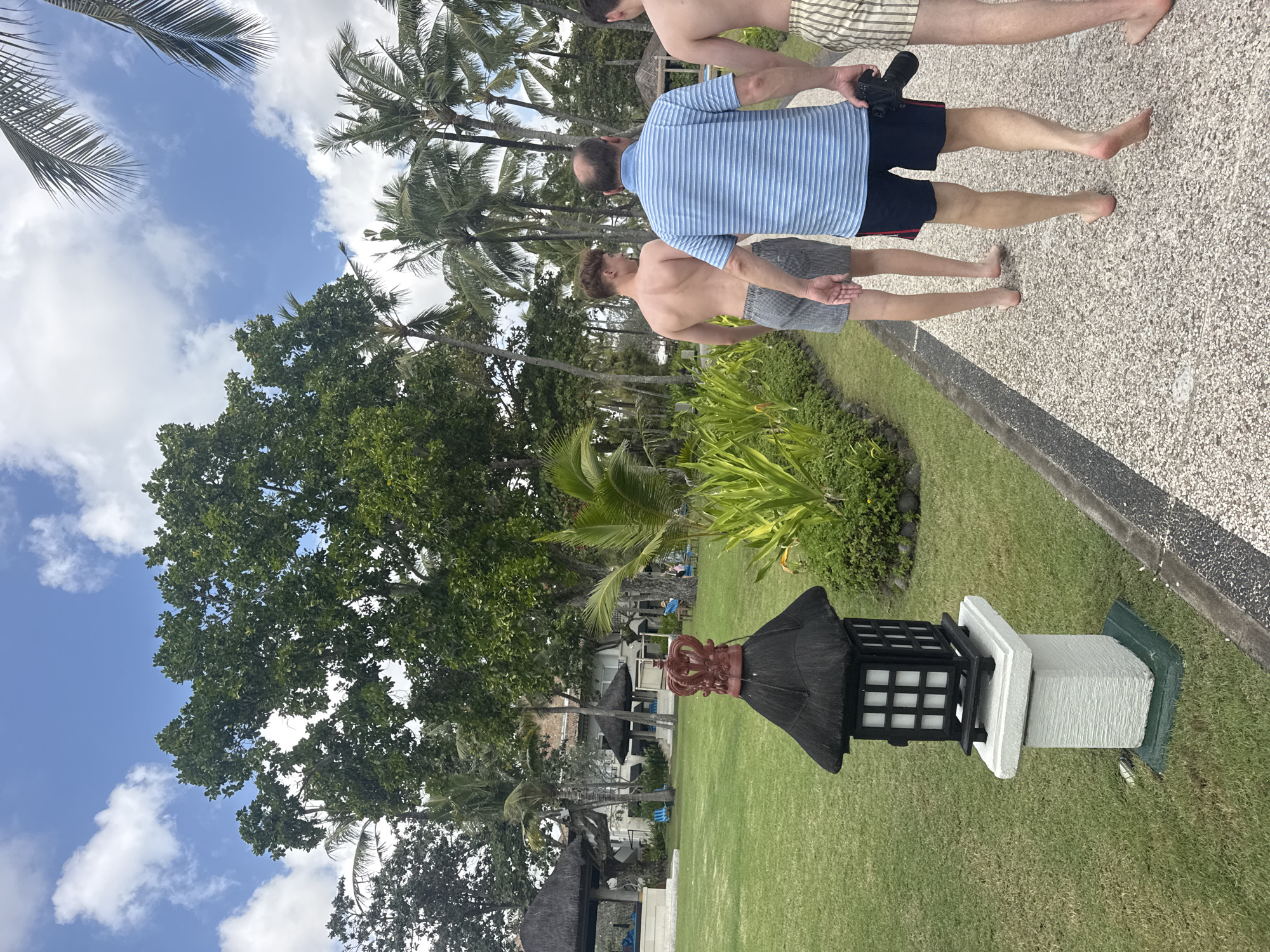 Three males are walking along a pebbled pathway in the city of Kuta Selatan, Indonesia. The two males in the foreground are barefoot, wearing swim trunks. The male on the left, wearing striped swim trunks, has his back to the camera. The male in the middle, wearing a blue and white striped polo shirt and dark swim trunks, is holding a black camera in his right hand. The third male, wearing striped swim trunks, is partially visible on the right. They are walking away from the camera, towards a lush, green landscape with palm trees and other vegetation. In the background, there is a building with a thatched roof and several open-air structures. The sky is blue with scattered white clouds. A decorative lamp post with a crown-shaped ornament is visible on the left side of the pathway. The area appears to be a resort or hotel.