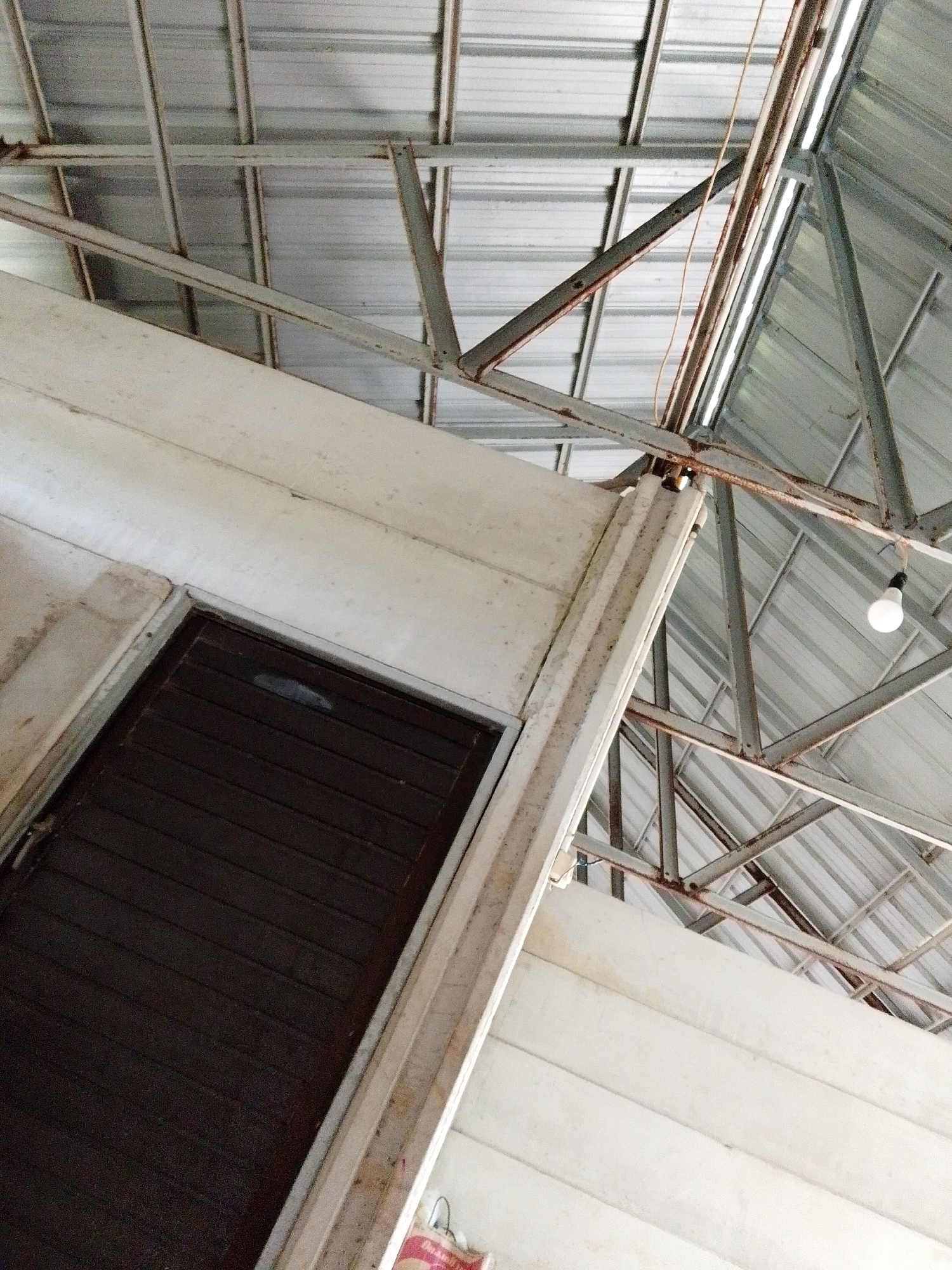 The image, taken in Bukit Ketok, Indonesia, shows an interior space. A closed, dark brown wooden door is in the lower left of the frame, set in a white frame. Above the door and along the right side of the frame is a white, slightly dirty wall. The ceiling structure is visible, constructed of metal beams and corrugated metal sheets. A single bare light bulb hangs from the ceiling on the right side. Rust is visible on some of the metal beams.