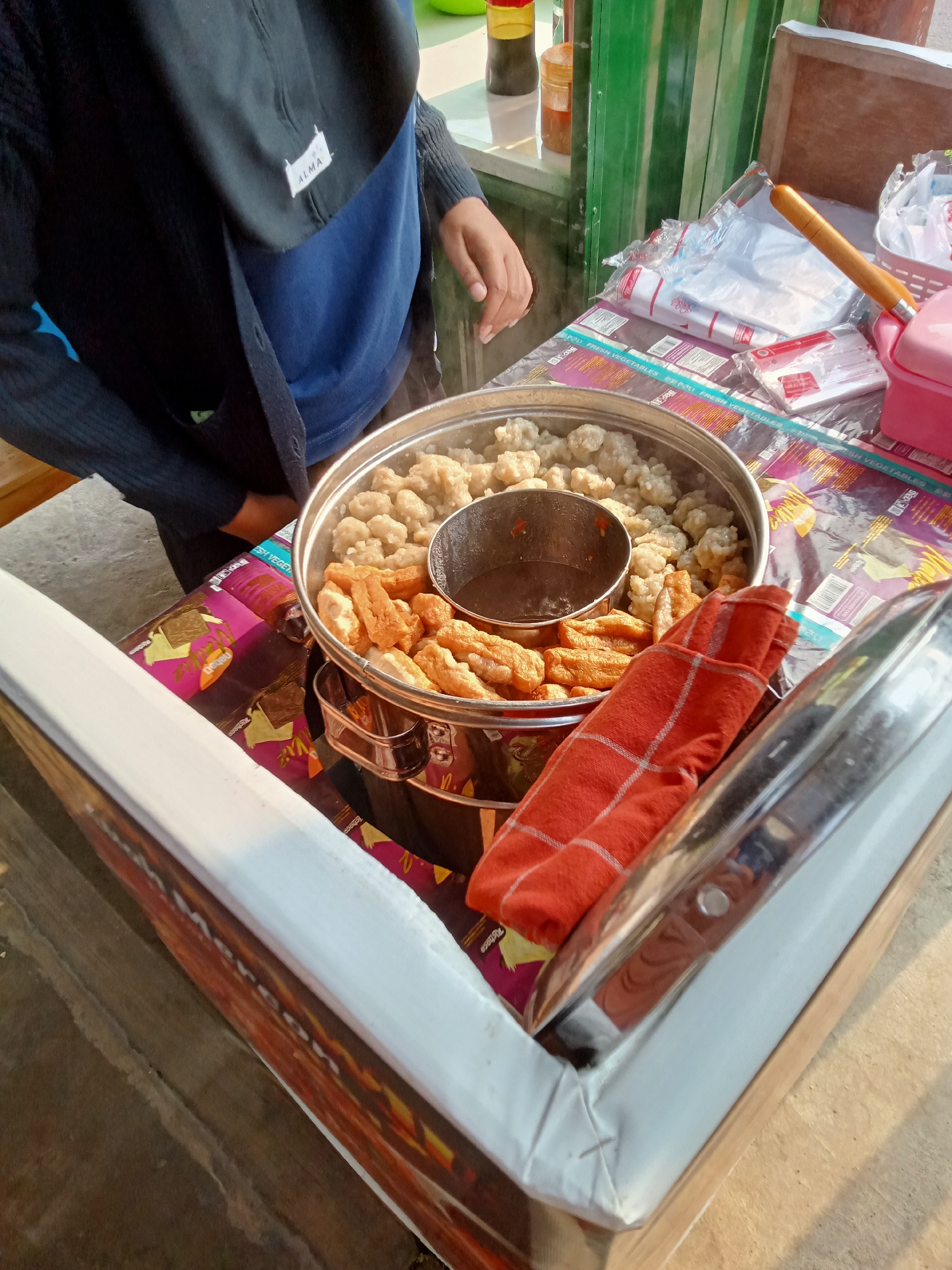 In Simo, Indonesia, a street food vendor's cart displays a steaming pot of Indonesian meatballs, locally known as "bakso," and fried tofu. The cart is covered with colorful packaging, and a hand, possibly belonging to the vendor, reaches towards the food. The scene is illuminated by natural light, suggesting it's daytime. The vendor wears a dark blue shirt and a black outer garment, with a white tag bearing the "ALMA" logo visible on the garment. The cart appears to be situated outdoors, possibly on a street or market.
