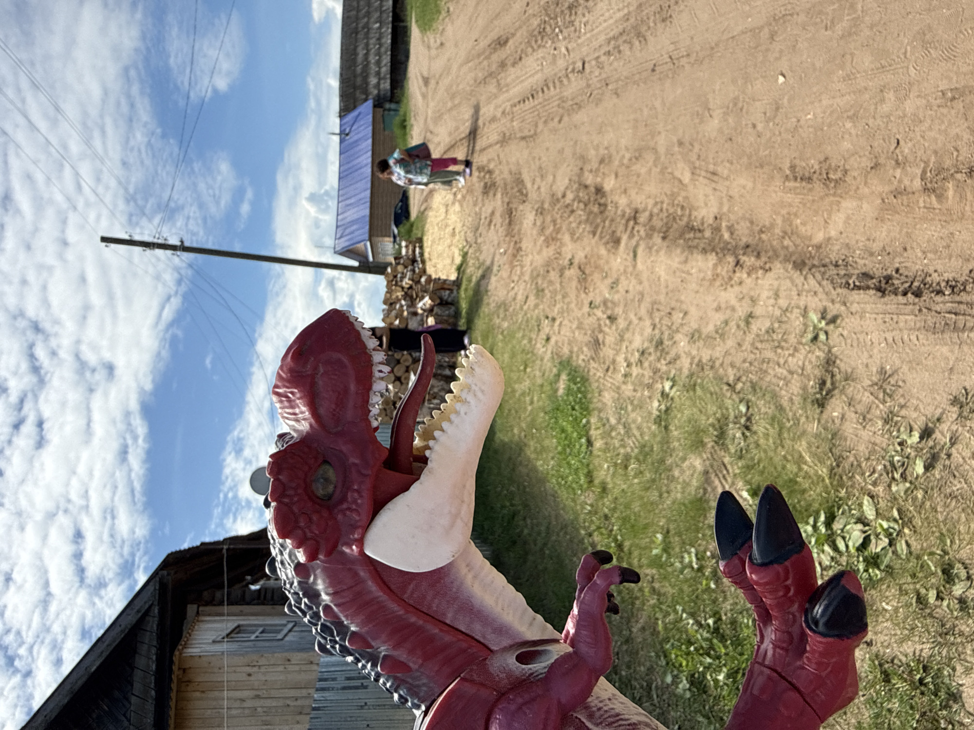 A large, red and black dinosaur toy, with its mouth open revealing white teeth and a red tongue, occupies the lower left foreground. Its textured skin and black claws are prominent. In the midground, a wooden house with a dark roof and a corrugated metal fence are visible behind the toy. A dirt road, marked with tire tracks, extends diagonally from the midground left to the background. Two individuals are walking away from the camera on the dirt road in the mid-to-background. One wears a light shirt and pants, carrying a bag. The other wears a patterned top and pink pants, also carrying a bag. A utility pole with power lines stands to the right of the first house. Further in the background, a house with a distinct blue roof and another traditional wooden house are visible. A pile of cut logs is stacked beside the road on the left. The scene is set under a blue sky with scattered white clouds, in a rural environment with sparse green vegetation along the roadside, located in Уральское, Russia.