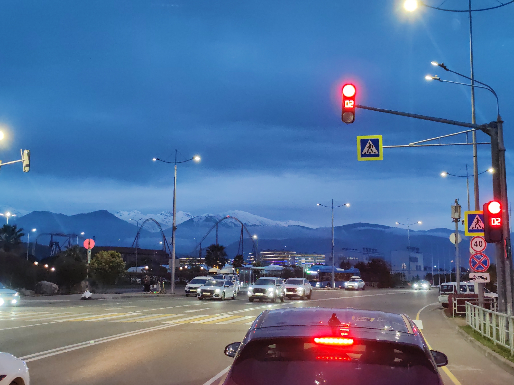 The image depicts an evening or twilight scene of a multi-lane road, captured from the perspective of a vehicle stopped at a red light. The sky is a deep blue, characteristic of dusk, with some lighter clouds visible. In the immediate foreground, the rear of a dark-colored car with illuminated red brake lights is prominent.
Ahead, several other cars are stopped at the intersection, their headlights on, casting light on the asphalt. A marked pedestrian crosswalk spans the road. To the right, a tall pole supports two red traffic lights, both displaying a digital countdown of "02". Below the main traffic light, a yellow square sign with a black pedestrian icon is affixed. Further down the pole, two circular road signs are visible: one white with a red border and the number "40" (likely indicating a speed limit), and another blue sign with a red diagonal line, typically signifying "no stopping" or "no waiting". Below this, a circular sign with a red border and a diagonal line over a tow truck symbol indicates "no towing" or "towing prohibited".
Streetlights line the road, emitting bright white and yellow light. In the mid-ground and background, behind the urban infrastructure, a range of large, rugged mountains, some appearing snow-capped, extends across the horizon. Between the road and the mountains, various buildings and a distinctive roller coaster structure from what appears to be an amusement park are visible, their outlines dark against the sky and illuminated by artificial lights. The overall scene portrays an active urban environment set against a dramatic natural landscape as night falls.
Location context: Unknown, Unknown.