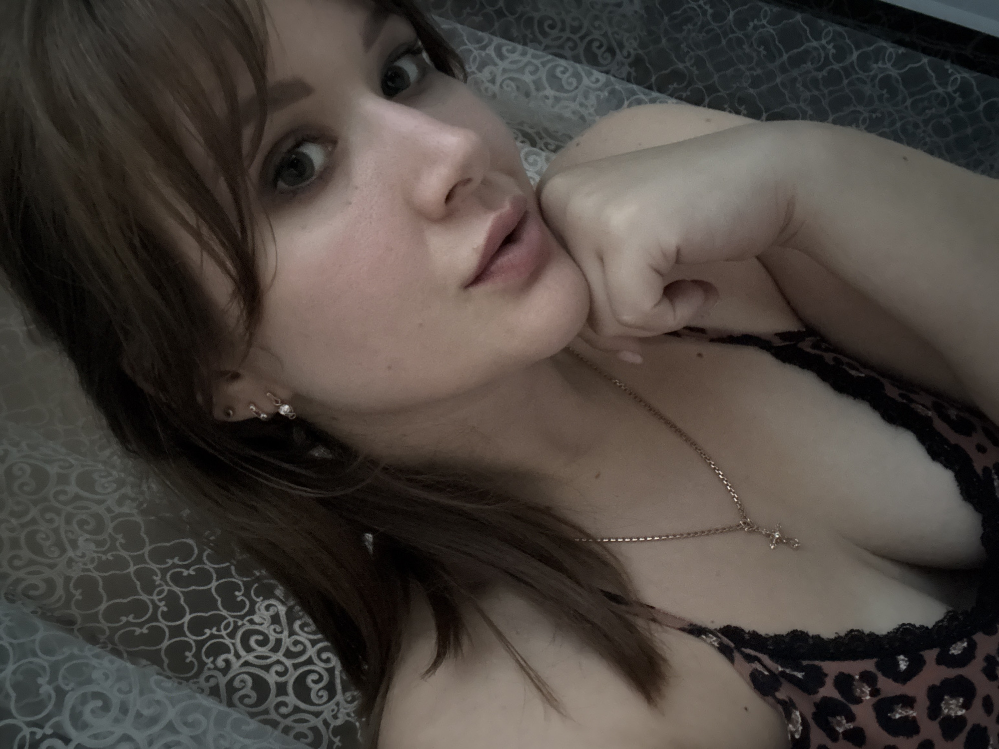 The media file is a close-up, self-taken photograph of a young woman lying down, likely in bed, given the bedding visible. She is looking towards the camera with a relaxed and contemplative expression, her head tilted and one hand resting on her chin. Her hair is a medium brown color and falls across her face, partially obscuring her left eye. She is wearing a leopard-print top with black lace trim, and a delicate silver necklace with a small cross pendant is visible around her neck. She has small hoop earrings in her ear. The lighting is soft and appears to be from an indoor source, suggesting it might be nighttime or a dimly lit room. The background consists of patterned bedding, adding to the intimate and personal feel of the image. There is no visible text in the image. The overall mood is one of quiet introspection and comfort.