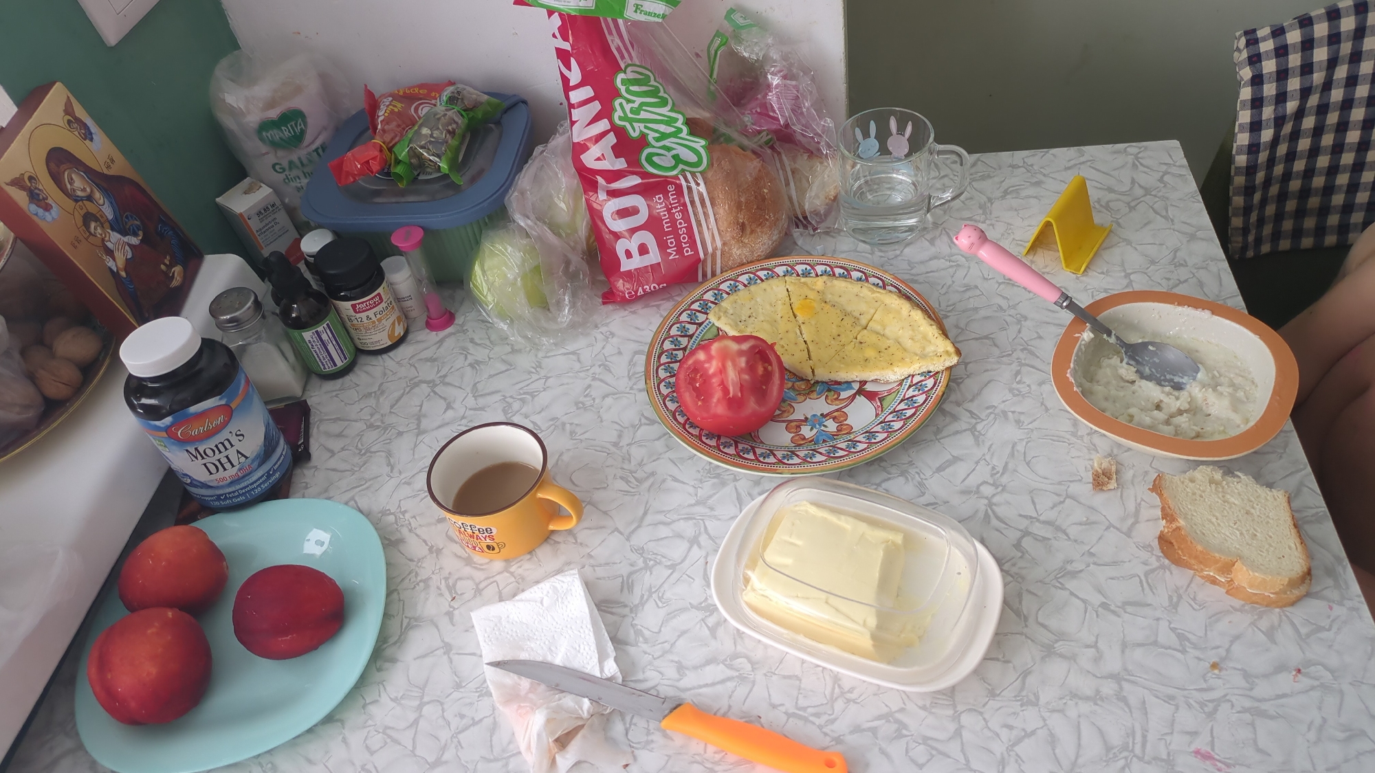 This is a top-down, medium shot of a table laden with breakfast items. The scene appears to be an indoor setting, possibly a kitchen or dining area. On the table, there's a plate with a tomato and a flatbread. Next to it, a bowl of what appears to be porridge with a spoon. To the left, a plate of peaches and a yellow mug with coffee. There are also bottles of supplements, a container of butter, and slices of bread. In the background, a checkered chair and part of a person's leg are visible, suggesting a person is seated at the table. The overall impression is a simple, home-cooked breakfast. No specific landmarks or elements in the image directly indicate the location as Chișinău, Moldova.