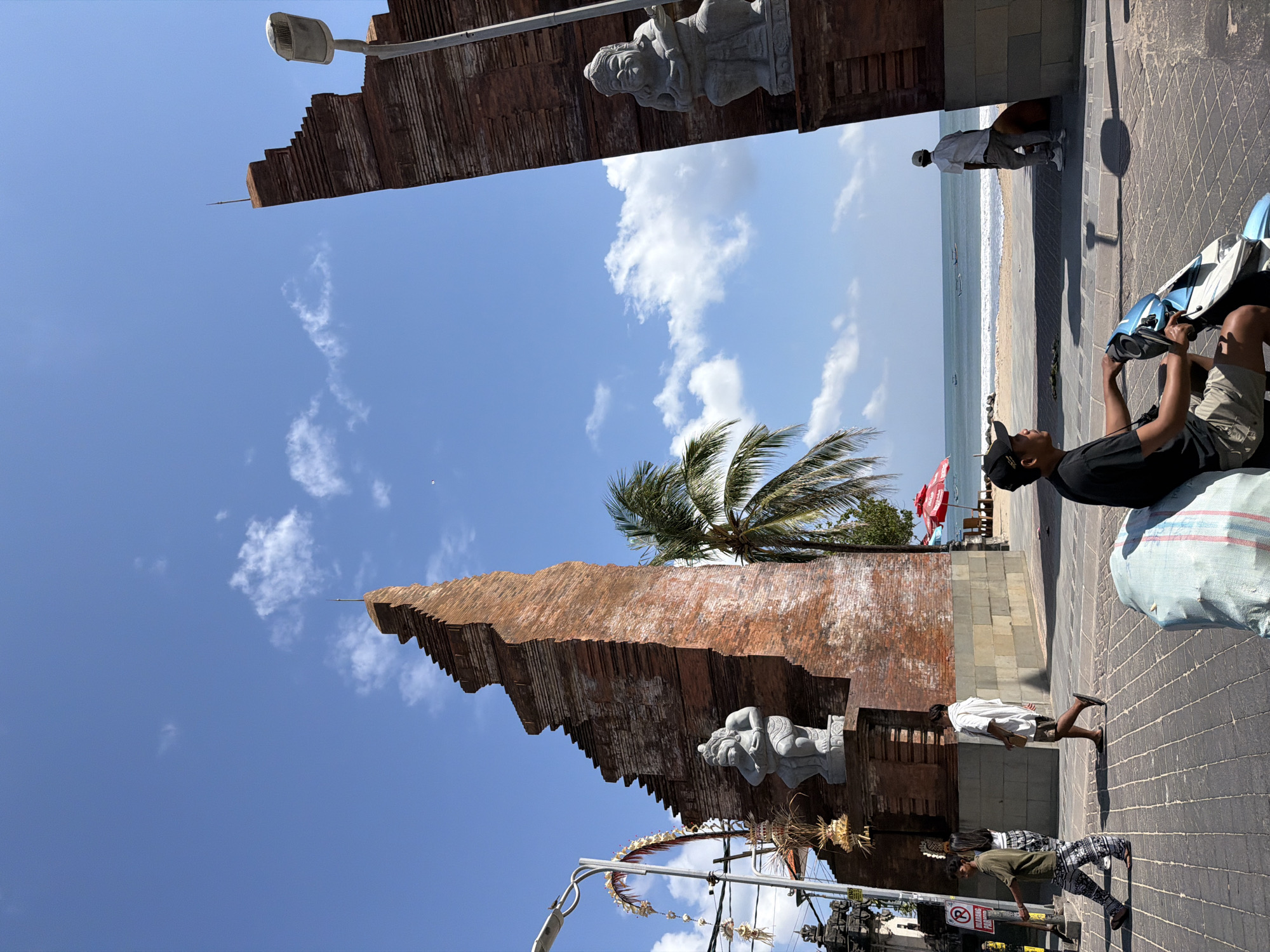 In Kuta, Indonesia, a wide, paved pedestrian walkway runs alongside the beach. The walkway is bordered by elaborate brick structures that appear to be temple gateways, each topped with a decorative finial and featuring stone statues of imposing figures. The gateway on the left is partially visible, while the one on the right frames a view of the ocean. A palm tree grows between the gateways, its fronds extending towards the sky.
Several people are present. On the left, a young man with dark hair wears a green t-shirt and patterned trousers, walking away from the camera. Beside him, a woman with dark hair wears a white top and patterned trousers, also walking away. Further to the right, a man in a white shirt and patterned shorts walks past the right gateway, carrying a small brown object. In the foreground, a man wearing a black t-shirt, a black baseball cap, and khaki shorts is seated on a light blue and white scooter, holding onto its handlebars. He faces away from the viewer, looking towards the sea. A large, light blue and white striped bag is placed on the ground near the scooter.
To the right of the gateway, a man in a white shirt and beige trousers stands near the beach, facing the ocean. A large brown bag or object rests on the ground beside him. In the background, the ocean stretches out to the horizon under a bright blue sky dotted with white clouds. The beach is visible, with several lounge chairs and a red umbrella set up. Small boats are scattered on the water. A lamppost stands to the right of the right gateway, its light fixture pointed downwards.