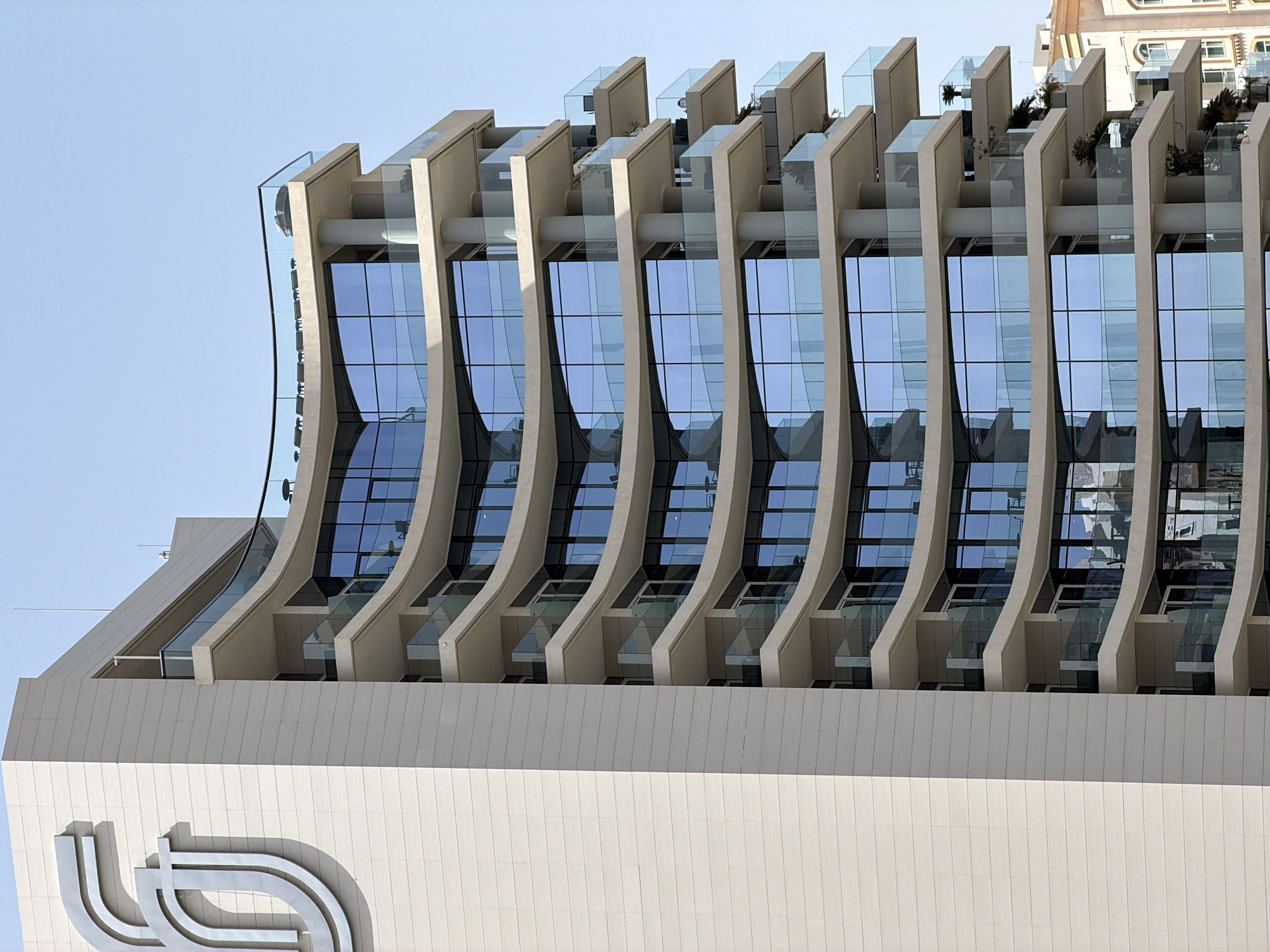A low-angle, full shot reveals a modern building in Lusail, Qatar. The building is characterized by its sleek design, featuring curved vertical beams that frame large glass windows, reflecting the sky. Small, elevated terraces with glass railings are visible above each window section, some containing small plants. The building's lower portion is clad in smooth, white panels with a stylized logo. The sky is clear and pale blue. Another building can be seen in the top right corner.