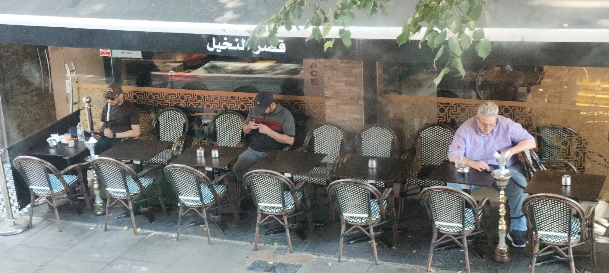 In an outdoor seating area of a cafe, three men are seated at separate tables. The man on the far left, wearing a dark t-shirt, baseball cap, and sunglasses, is smoking a shisha pipe and looking at his phone. He is seated at a dark rectangular table. To his right, another man in a grey and red t-shirt and baseball cap is also looking at his phone, seated at a similar table. The man on the far right, wearing a light purple shirt and blue jeans, is also smoking a shisha pipe and is seated at a dark rectangular table. He has a bottle of water on the table next to him. All three tables are accompanied by wicker chairs with patterned cushions. The cafe has a dark facade with Arabic script above the windows, and decorative patterned screens behind the seating area. The ground appears to be paved with concrete. The overall scene suggests a relaxed atmosphere in an urban setting.