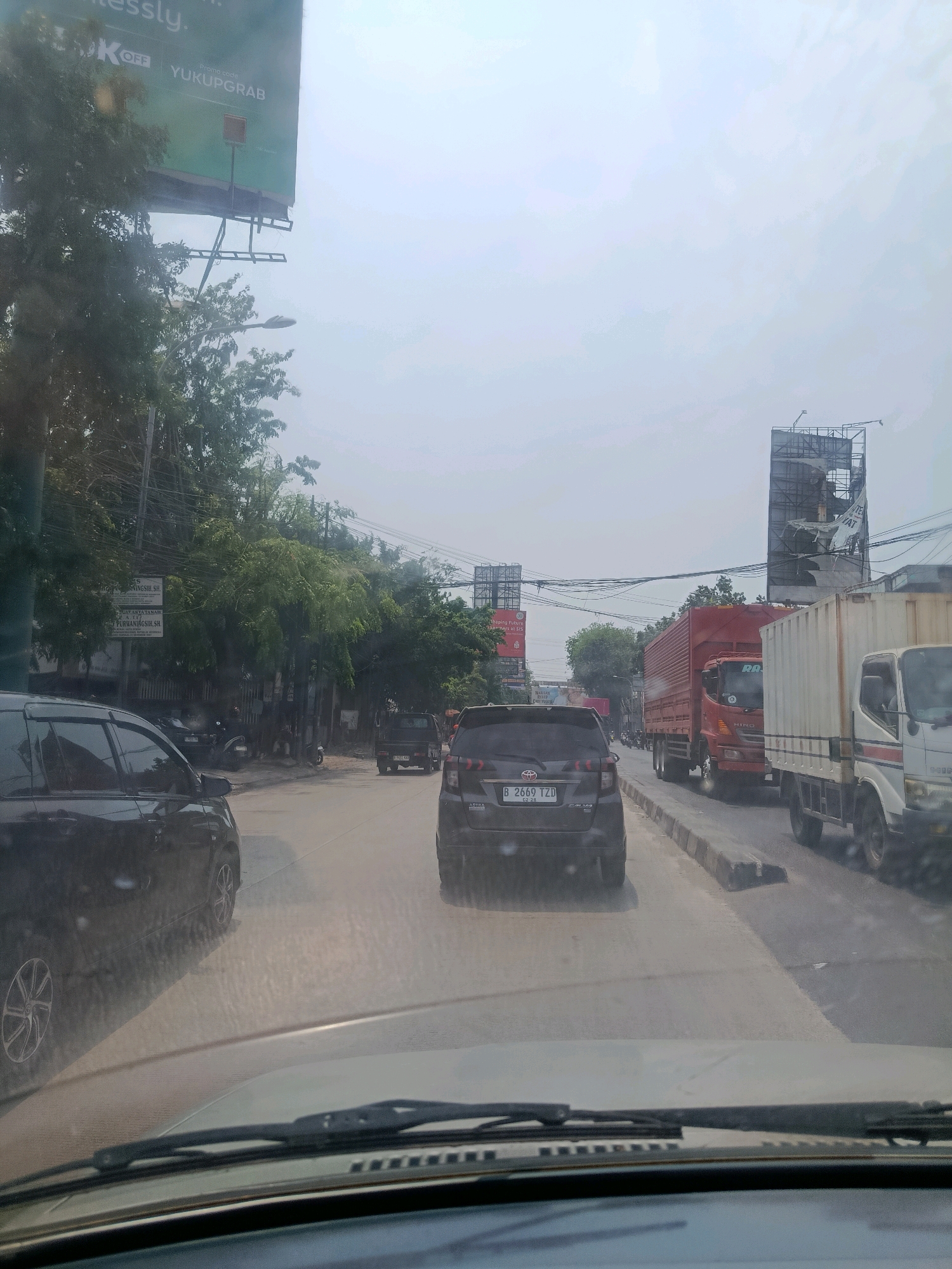 The image depicts a street scene in Bekasi, Indonesia, during daylight. The perspective is from inside a vehicle, looking forward. Traffic is moderate, with several cars and trucks visible on the road. In the foreground, the hood and windshield wipers of the vehicle are prominent. A black SUV is to the left, and a darker vehicle is in front. To the right, two trucks are in the lanes, one red and one white. A tall billboard structure is visible in the background, partially obscured by trees and overhead wires. The sky is overcast with a hazy, bright light. The overall environment suggests a typical urban roadway with commercial signage and infrastructure.
