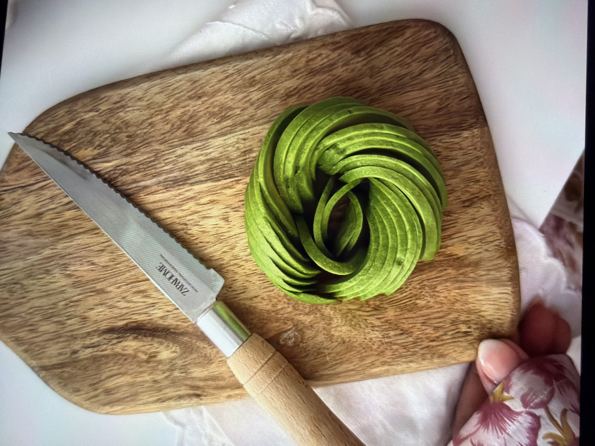 This overhead close-up image captures a culinary arrangement on a wooden cutting board. The main subjects are a knife, a spiral-shaped avocado, and various fabrics.
On the left, a knife with a serrated, silver-colored blade and a light wooden handle rests on the board. The brand "ZARA HOME" is visibly engraved on the blade, followed by smaller, indistinct text.
To the right of the knife, a bright green avocado has been skillfully sliced into thin strips and meticulously arranged into a concentric spiral, commonly known as an "avocado rose." The vibrant green of the avocado stands out against the natural grain of the wooden cutting board. The board itself is light-toned, with visible wood grain and a rounded, organic shape.
The cutting board is placed on a white or light-colored, textured fabric, possibly a tablecloth or napkin. On the far right, a human hand with light-colored fingernail polish is visible, holding a piece of fabric adorned with a floral pattern featuring pink, red, and yellow motifs on a light background.
The scene appears to be indoors, under even ambient lighting, suggesting food preparation or presentation. No specific time of day or location cues for Ros Na Rí, Ireland are discernible from the visual information within the image. The overall impression is one of artful food styling.