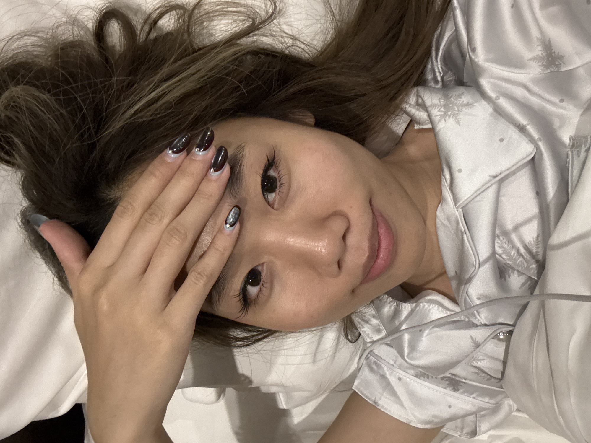 A person is lying on a white pillow, wearing a light-colored pajama top patterned with snowflakes. They have brown hair with blonde highlights and are touching their forehead with their left hand, displaying nails painted dark with light accents. The location is unknown and cannot be determined from the image.