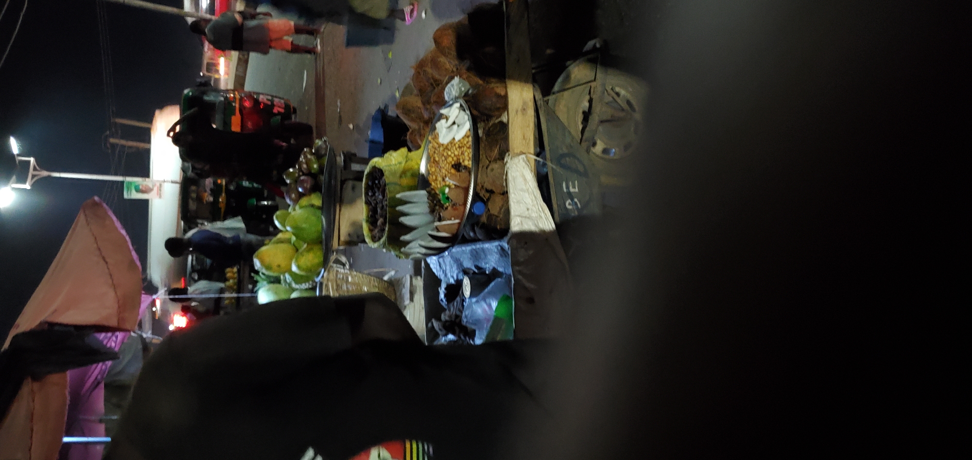 The image depicts a nighttime scene in an urban environment. A street food vendor is present, with displays of mangoes, coconuts, and snacks. A black object is in the foreground, obstructing the view of the central portion of the image. Street lights are visible. Several auto rickshaws are parked. Pedestrians are visible in the background.