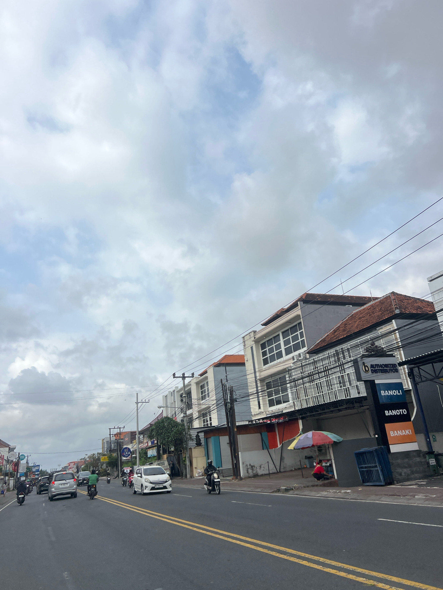 The image depicts a street scene in Denpasar, Indonesia, characterized by a moderate amount of traffic, primarily consisting of motorcycles and cars. The sky is overcast with a mix of white and gray clouds. The road is asphalt with double yellow lines marking the lanes. Buildings line the right side of the road, exhibiting a mix of modern architecture with glass facades and tiled roofs, typical of commercial or residential structures in the region.
Several businesses are visible, with signage including "AUTHORIZED DISTRIBUTOR," "BANOLI," "BANOTO," and "BANAKI." A small stall with a colorful umbrella is set up on the sidewalk in front of one of the buildings, and a person in a red shirt is seated nearby. Other elements in the scene include utility poles with electrical wires, trees, and a person on a motorcycle wearing a black helmet. The overall impression is of a busy urban environment during daylight hours.