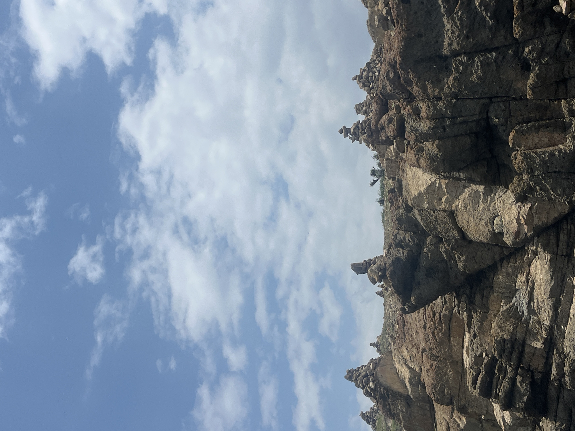 The image depicts a rocky coastline with numerous cairns (piles of stones) stacked on top of the cliffs. The sky above is blue with scattered white clouds. The terrain is rugged and appears to be arid or semi-arid. The stacked stones suggest a human presence and possibly a tradition or practice of marking the landscape. The location is identified as Mui Ne, Vietnam. The scene is devoid of people and any visible actions are limited to the static presence of the rocks and cairns. The environment is characterized by natural rock formations and the sky.