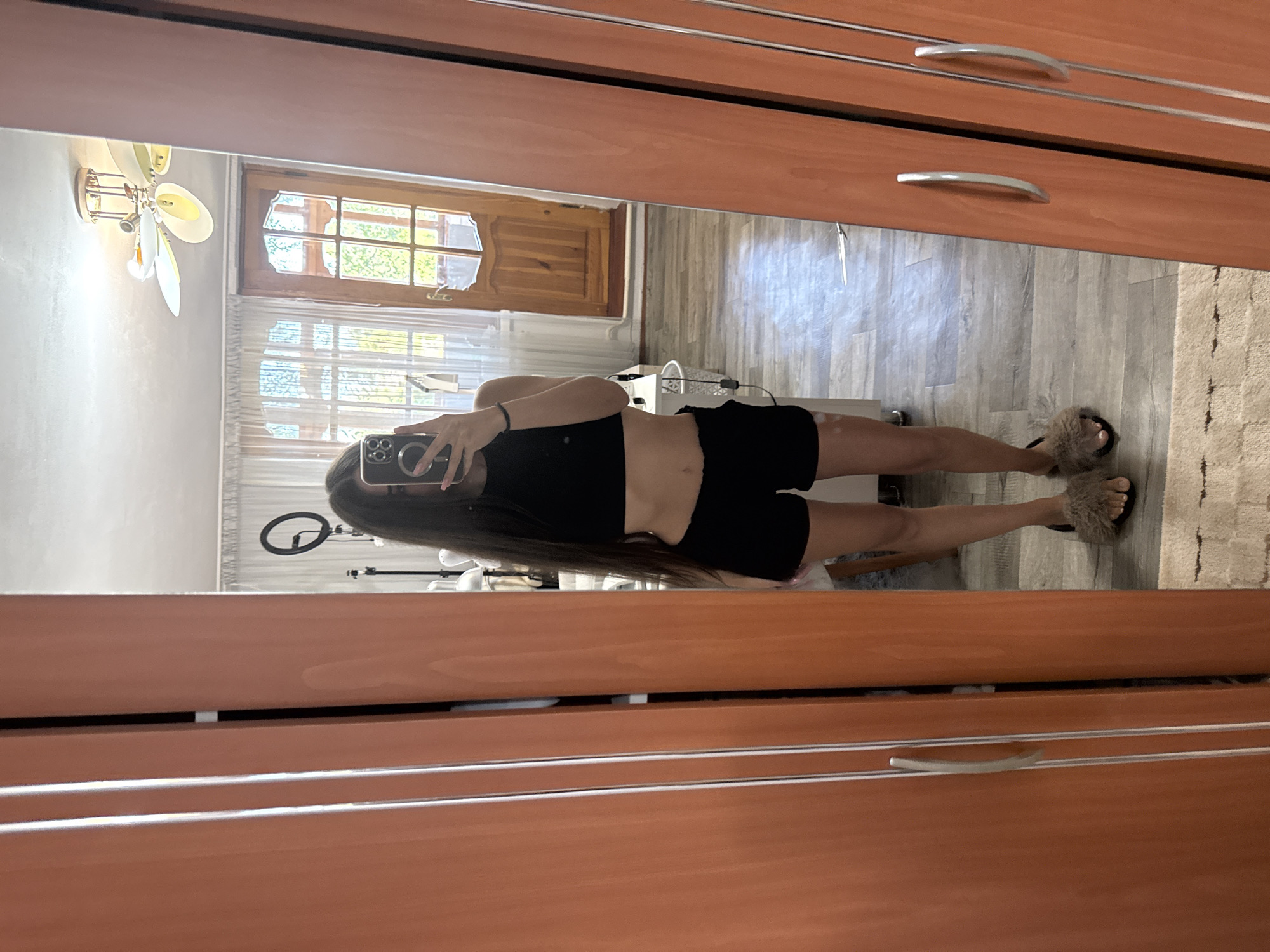 A young woman stands in a room, holding a phone to take a mirror selfie. She is wearing a black crop top and black shorts. She has long dark hair and is wearing furry slippers. The room appears to be a bedroom, with a wooden door and windows visible in the reflection of the mirror. The floor is covered with a light-colored laminate, and a rug is partially visible. A ceiling fan is mounted on the ceiling. The overall setting suggests a casual, domestic environment, possibly in Basarabeasca, Moldova.