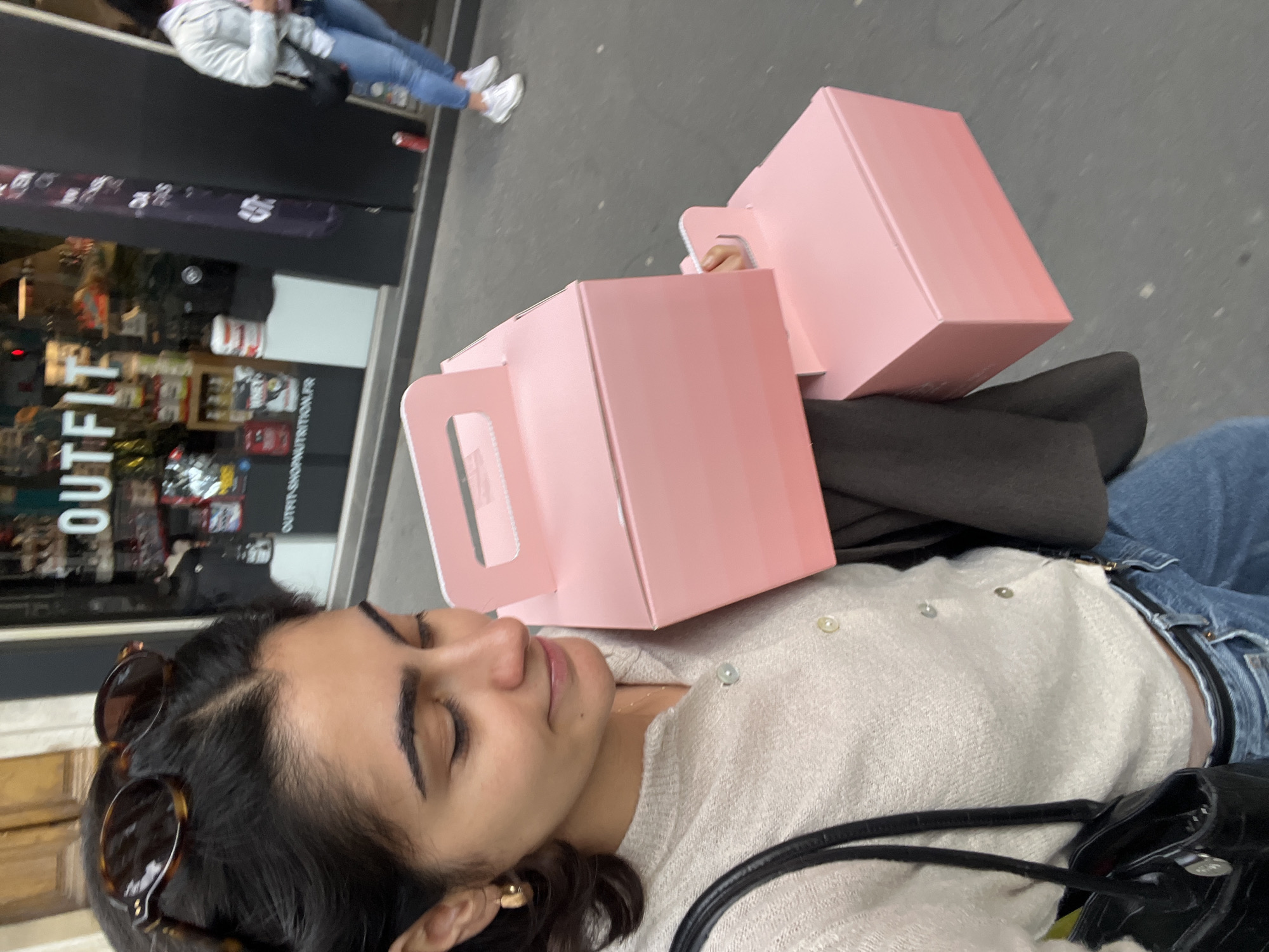 A medium-shot, canted, outdoor photograph captures a woman with brown hair wearing jeans and a cream-colored, button-down sweater, positioned on a street, likely in Paris, France. She is holding two pink boxes with handle cutouts. She has her eyes closed and is holding the boxes on her chest. Sunglasses are pushed up on her head. Her accessories include a gold earring, and a black bag. The environment includes an "OUTFIT" shop with reflected images, a gray sidewalk, and other pedestrians.