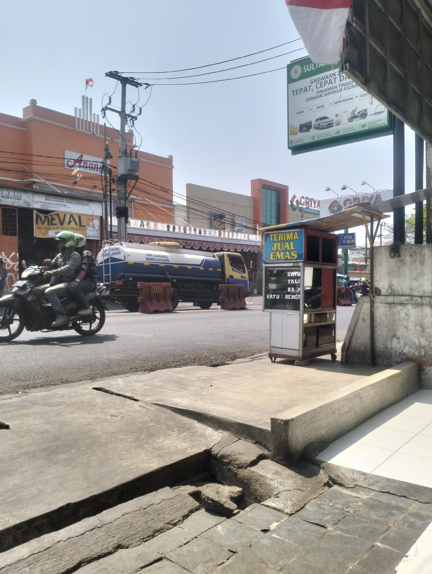 On a bright, clear day, a motorcycle carrying two individuals travels down a street in Kel. Pasir Jati, Indonesia. The rider, wearing a green helmet, guides the motorcycle while the passenger is seated behind. To the right of the road, a small street vendor stall displays signs advertising the sale of gold. Further down the street, a large water tank truck, labeled "AIR GUNUNG MANGILAYANG," is parked, with orange traffic barriers on either side. In the background, buildings with orange and white facades are visible, along with overhead power lines and utility poles. The foreground shows a cracked concrete sidewalk and paved road.