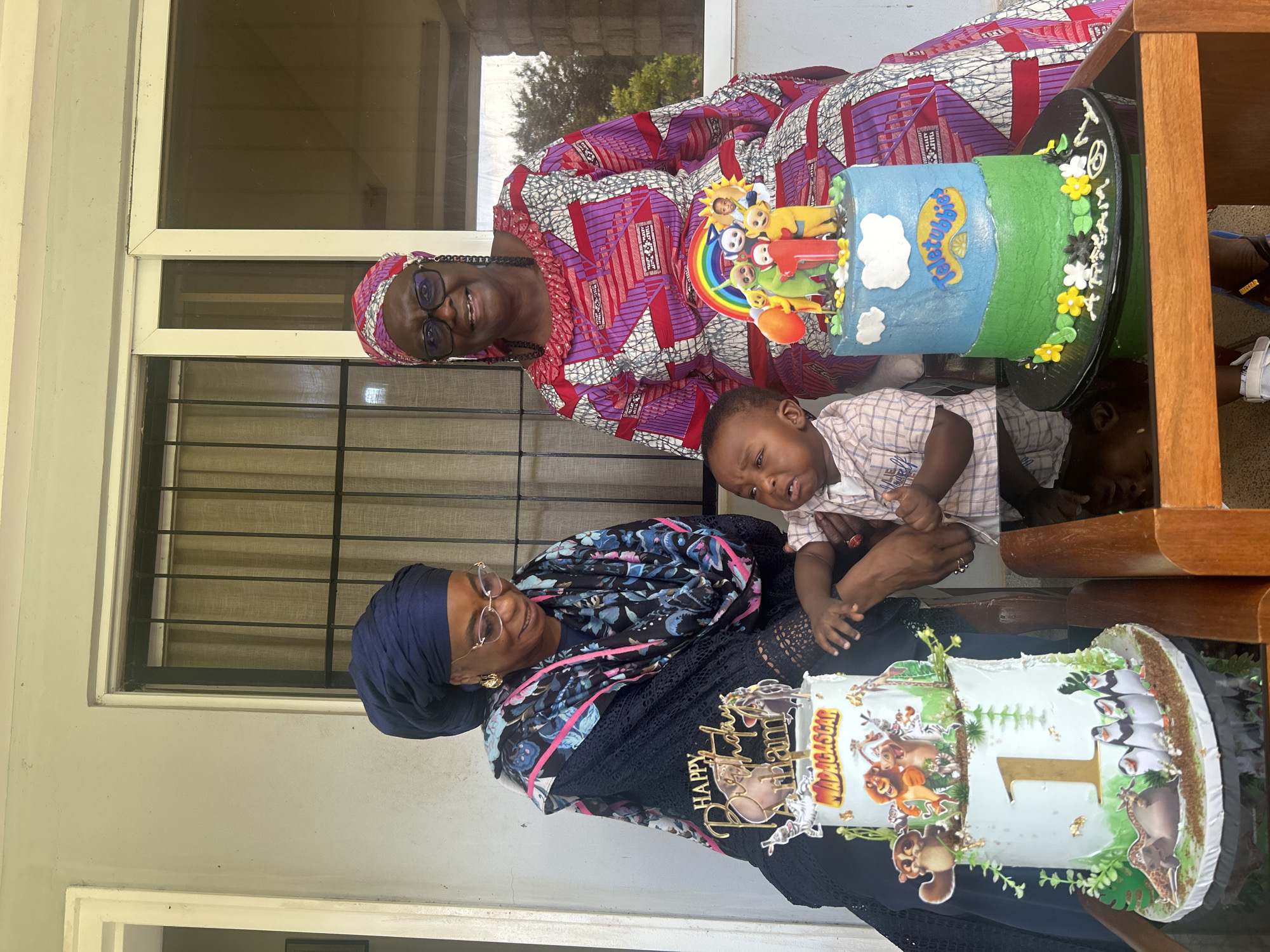 In Dutsen Kato, Nigeria, on a covered outdoor patio, two women and a baby are gathered around birthday cakes. The woman on the left wears a dark blue headscarf and a black and blue patterned dress. She has glasses and is looking down at a two-tiered cake adorned with Madagascar characters and a large '1'. The woman on the right wears a pink and purple patterned dress and a matching headscarf. She also wears glasses and smiles at the camera. Between the two women, a baby boy in a plaid shirt is being held by one of the women. He looks upset. In front of the woman on the right is a second cake decorated with Teletubbies and a rainbow. The cakes are placed on a reflective surface, possibly a table. Behind the group, a window with vertical blinds and a white frame is visible, suggesting the patio is attached to a building. The lighting indicates it is daytime.