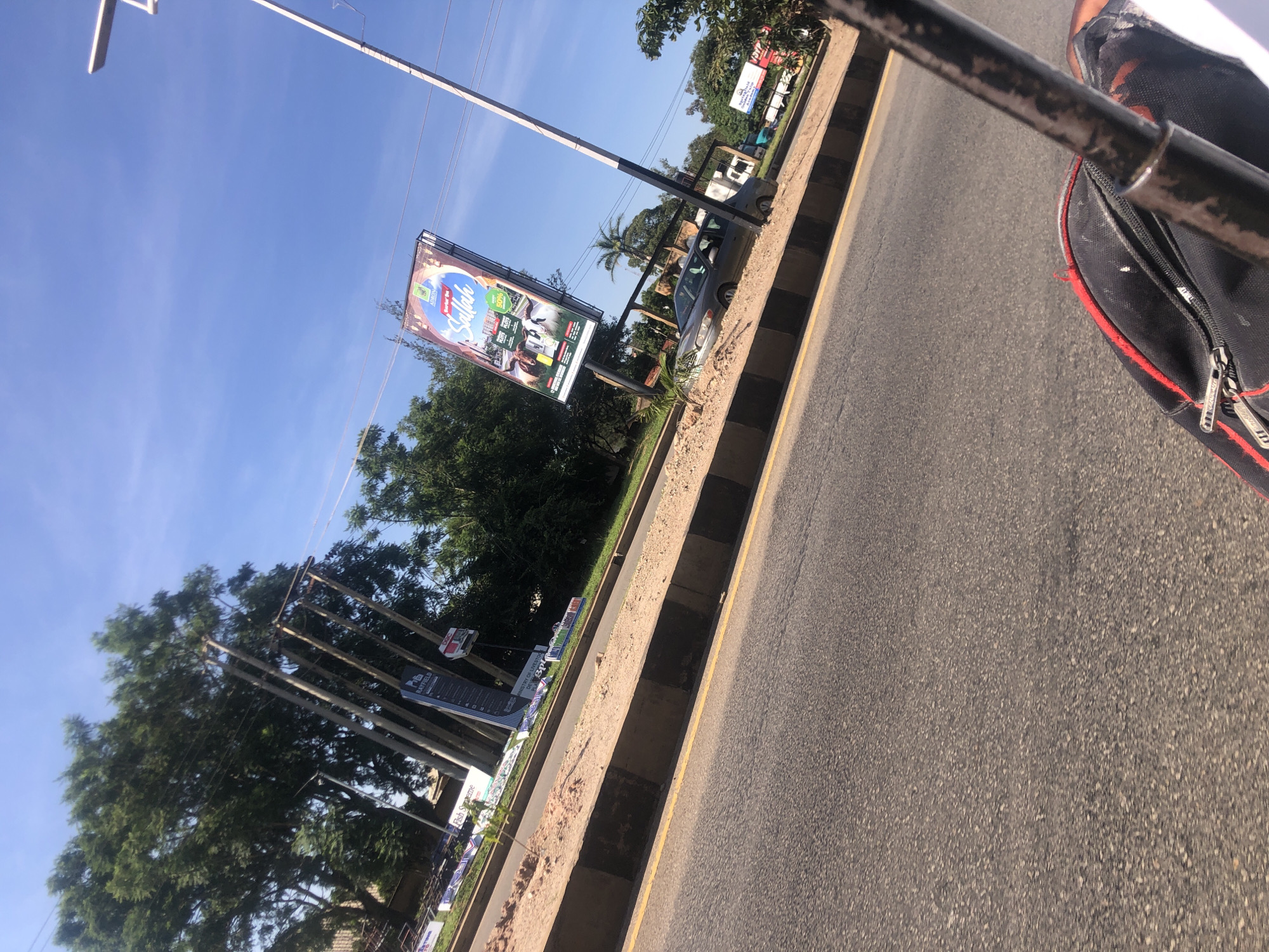 A tilted, eye-level, full shot captures a busy street scene in Jos, Nigeria. A gray sedan is parked on a dirt median on the right side of the road, partially obscured by a large billboard advertising "Sallah." The billboard is colorful, featuring images of livestock and green fields. Tall utility poles and lush green trees line the background. The road itself is paved and shows signs of recent construction, with a dirt berm and concrete barriers separating it from the median. A yellow line marks the edge of the road. In the foreground, a black bag with red trim and a metal pole are visible, suggesting the photo was taken from a moving vehicle. The sky is a clear, bright blue.