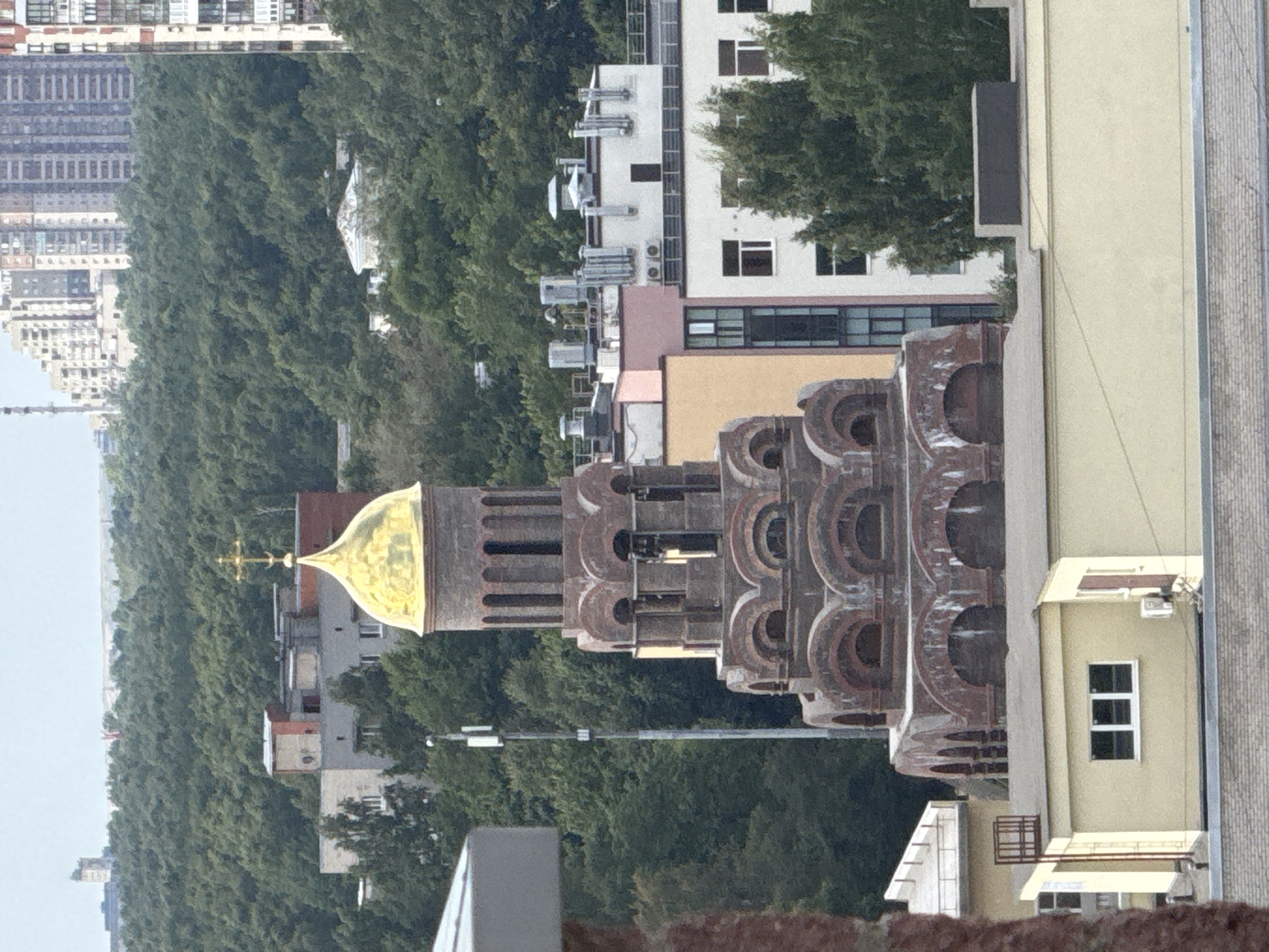 The image shows an Eastern Orthodox church with a golden dome and a cross at the very top. The church is made of brick and has multiple levels with arched windows. It is surrounded by trees and other buildings. In the background, there are tall apartment buildings. A cell tower is visible to the left of the church. The scene appears to be in an urban area, possibly a residential neighborhood, with a mix of old and new architecture. The lighting suggests it is daytime, possibly overcast.